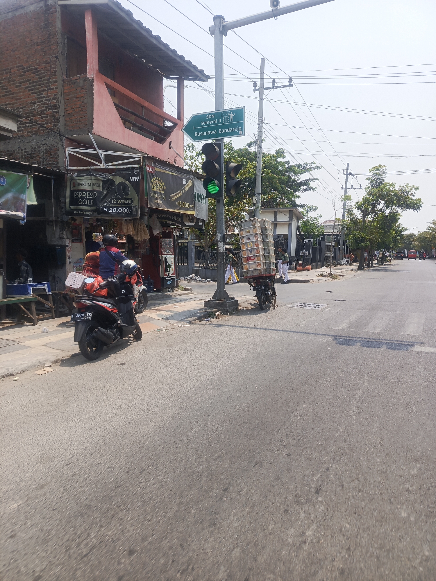 The image shows a street scene in Surabaya, Indonesia, likely on a sunny day with clear skies. On the left side of the street, there is a two-story building with exposed brick on the upper level and a pink facade on the lower level. Various signs are displayed on the building, including one for "SDN Sememi II" (a school) and another pointing towards "Rusunawa Bandarejo" (a housing complex).
In front of the building, there is a traffic light which is currently green. Several people and vehicles are present. Two motorcycles are parked on the sidewalk, and one of them is being ridden by a person wearing a blue jacket and an orange headscarf. Another motorcycle is parked further down the street. In the middle of the road, a motorcycle is loaded with many plastic crates, possibly for delivery. Several people, including some in what appear to be school uniforms, are walking on the sidewalk. Further down the street, cars and another motorcycle are visible. The road surface is asphalt, and a pedestrian crossing is marked. The overall environment suggests a commercial or residential area with moderate traffic.
