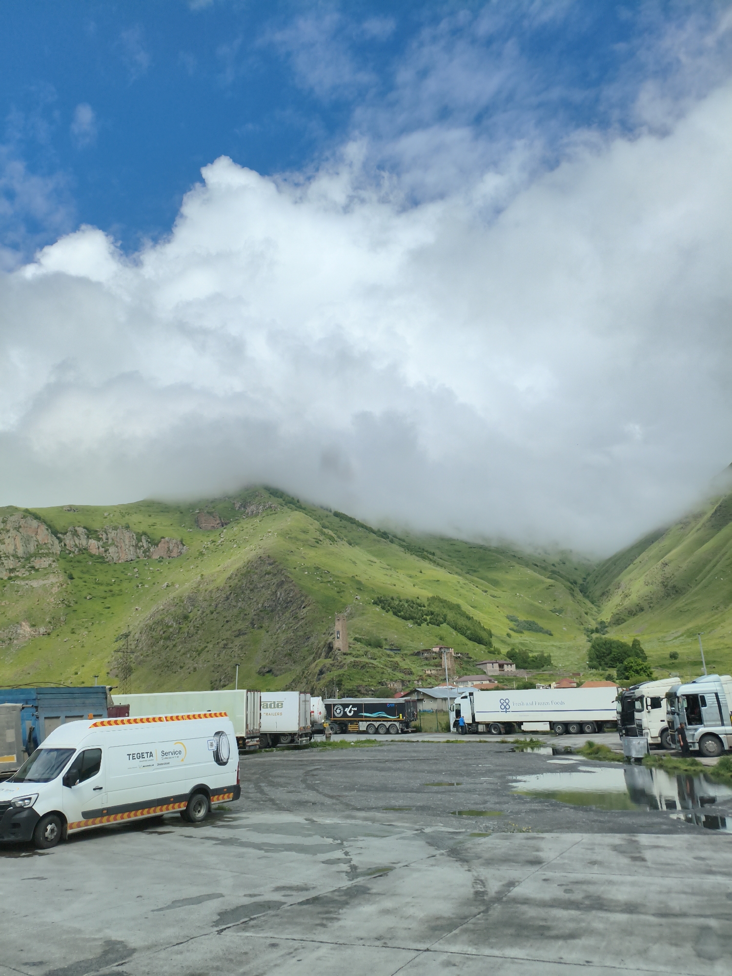 A white van with "TEGETA Service" branding and reflective orange and yellow stripes is parked on a wet asphalt lot. Several large trucks, including a white truck with "Fresh and Frozen Foods" on its side and another with "Trade Trailers" on its side, are parked behind it. In the background, green, rocky mountains are partially obscured by low-hanging clouds. A small, stone tower and scattered buildings are visible on the mountainside. The sky is blue with scattered white clouds. The scene appears to be a rest stop or service area in a mountainous region.