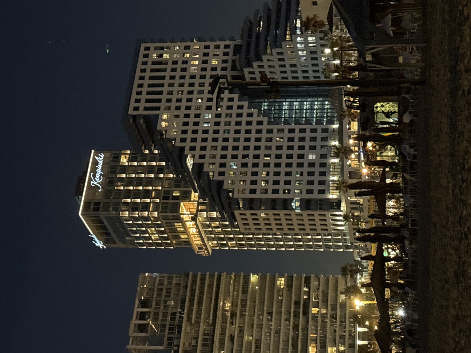 The image depicts a nighttime scene on a beach in Tel Aviv, Israel, featuring the Kempinski Hotel and another large modern building. The beach itself is dark, with the sand visible in the foreground. Several beach umbrellas and lounge chairs are set up on the sand, some appearing to be part of an outdoor seating area for a restaurant or bar. Palm trees are scattered across the background, illuminated by artificial lights.
The Kempinski Hotel is a tall, modern building with illuminated windows and a glowing sign at the top. Adjacent to it is another white, tiered building with a grid-like pattern of windows. The overall impression is one of urban nightlife near the coast, with the city lights contrasting with the dark sky. There are no discernible people or interactions visible in the foreground or on the beach. A faint green light source, possibly an aircraft or drone, is visible in the dark sky above the buildings.