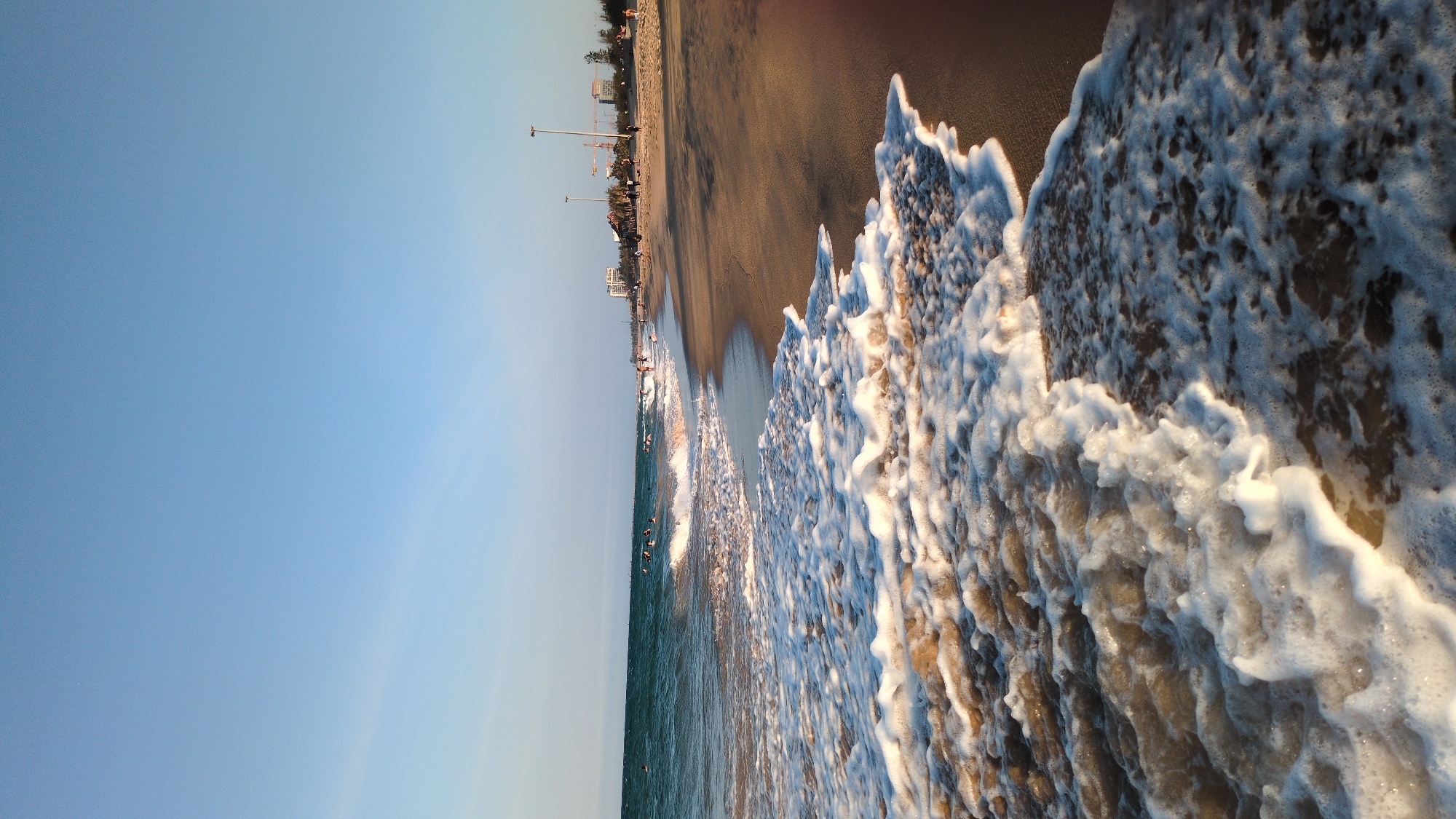 The image shows a beach in Thành phố Tuy Hòa, Vietnam, viewed from a ground-level, sideways perspective. The scene is divided into thirds: a blue sky on the left, a coastline/beach in the middle, and frothy waves occupying the right. The sky is predominantly clear blue, transitioning to a lighter hue near the horizon. The coastline features a dark brown sandy beach, with sparse buildings, light posts and cranes visible. Along the water’s edge, there are people swimming in the ocean. The water transitions from a dark green/blue further out to a lighter turquoise closer to the shore. The foreground is dominated by the white foam and texture of ocean waves washing ashore.