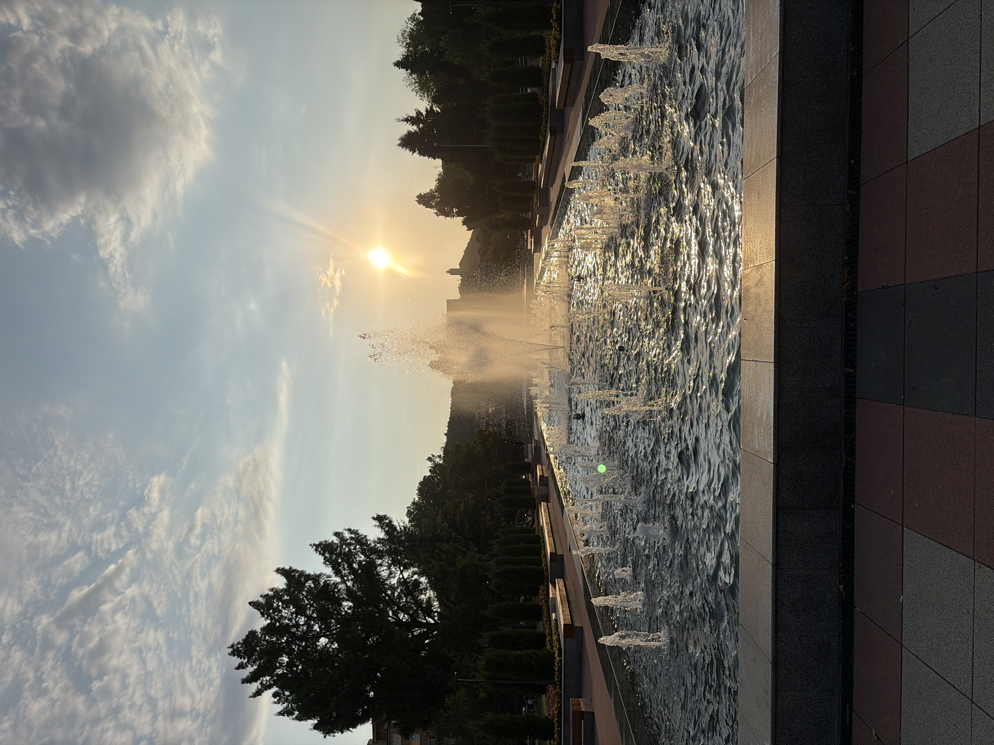 The image depicts a large, multi-tiered fountain with numerous jets of water shooting upwards, creating a dynamic and sparkling effect. The sun is setting or rising in the background, casting a warm, golden light that reflects off the water's surface and creates a sunbeam effect in the sky. The fountain is situated in what appears to be a park or public square, with manicured trees and bushes lining the edges of the water. In the distance, a building or monument is visible on a hilltop. The foreground shows a paved area with a patterned design. This scene is located in Montana, Bulgaria, specifically at the main fountain in the city center, a popular spot for recreation and gatherings.