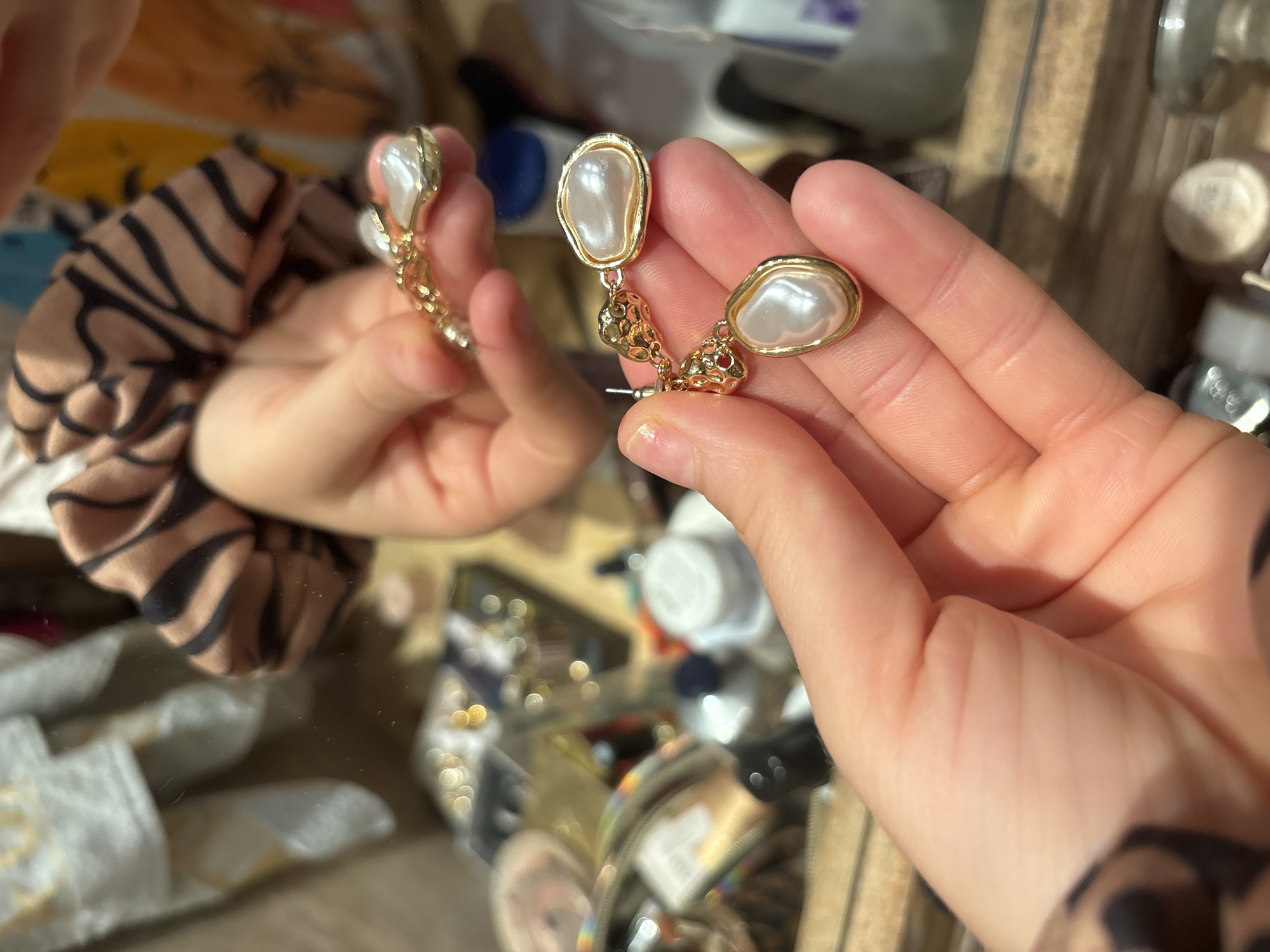 A person holds up two gold earrings with pearl-like centers. The person is likely looking in a mirror, as their reflected hand holds the other earring. The background appears to be a cluttered surface, possibly a vanity, with various cosmetic or personal care items visible. The reflection suggests this is indoors and potentially a personal space. The location in Budva, Montenegro, is not discernible from the provided visual information.