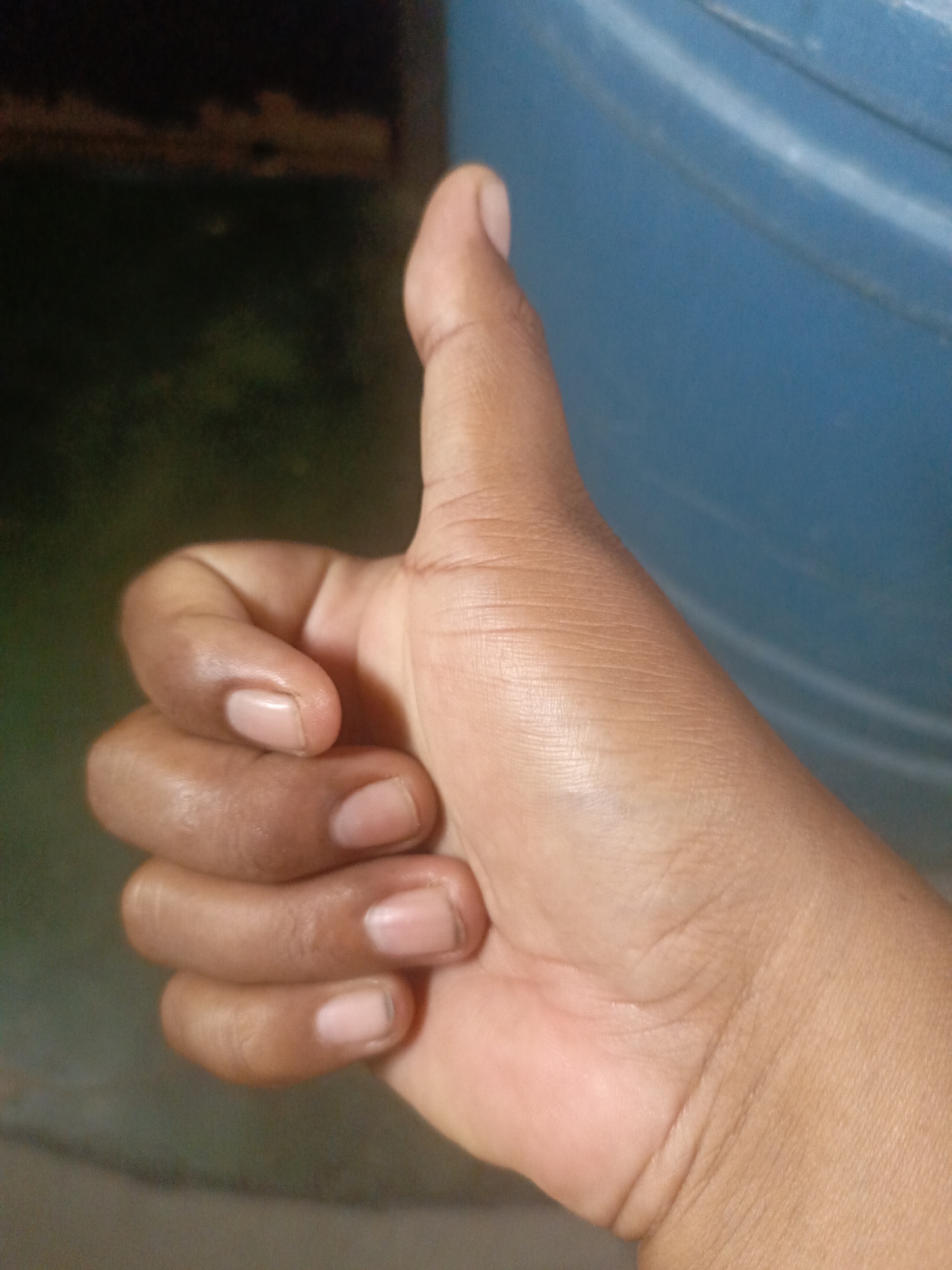 A close-up shot of a dark-skinned hand giving a thumbs-up gesture. The hand is positioned against a blurry background of dark and light areas and a light blue, potentially plastic, surface. No specific details suggest a location in Juan Griego, Venezuela.