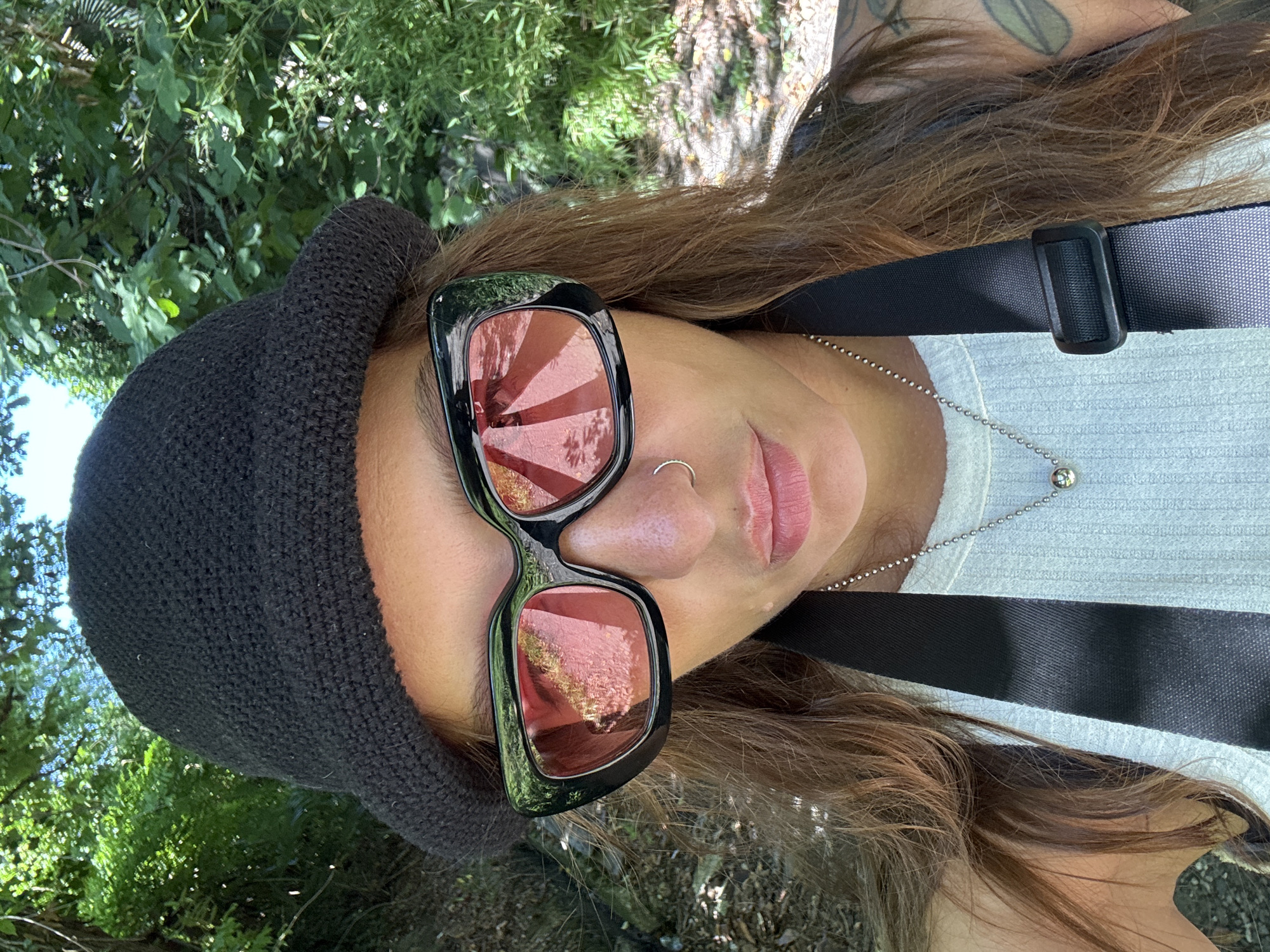 A person is taking a selfie outdoors, with lush green foliage and trees in the background, suggesting a park or natural area. The person is wearing a black bucket hat, large black sunglasses with pink lenses, and a white ribbed tank top. A black strap, possibly from a camera bag, is visible across their chest. The person has long brown hair and a silver necklace with a small pendant. The overall lighting suggests it is daytime. The scene is likely in Sochi, Russia, known for its abundance of natural beauty and parks.