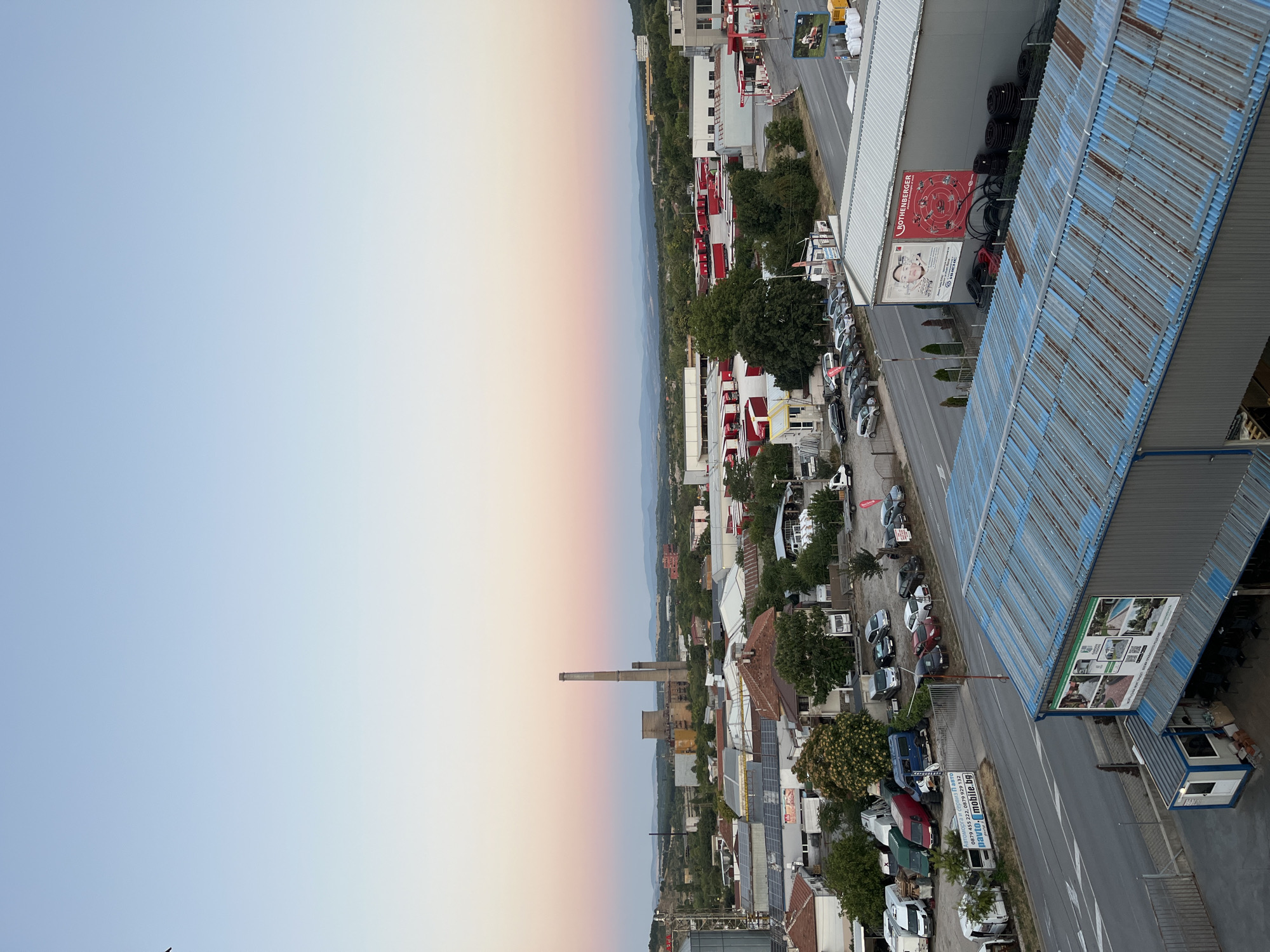 An aerial, wide-angle, eye-level shot captures an industrial and residential area at dusk in Vratsa, Bulgaria. The sky displays a soft gradient of pink and light blue. In the distance, a tall, industrial smokestack rises above a cluster of buildings, some with solar panels on their roofs. A main road runs horizontally through the mid-ground, lined with parked cars and trucks. Several commercial buildings are visible, including one with a large "ROTHENBERGER" advertisement. A corrugated metal roof dominates the foreground, with a small, white-and-blue modular building situated to its left. The overall scene suggests a functional, working environment with a blend of industrial and residential elements.