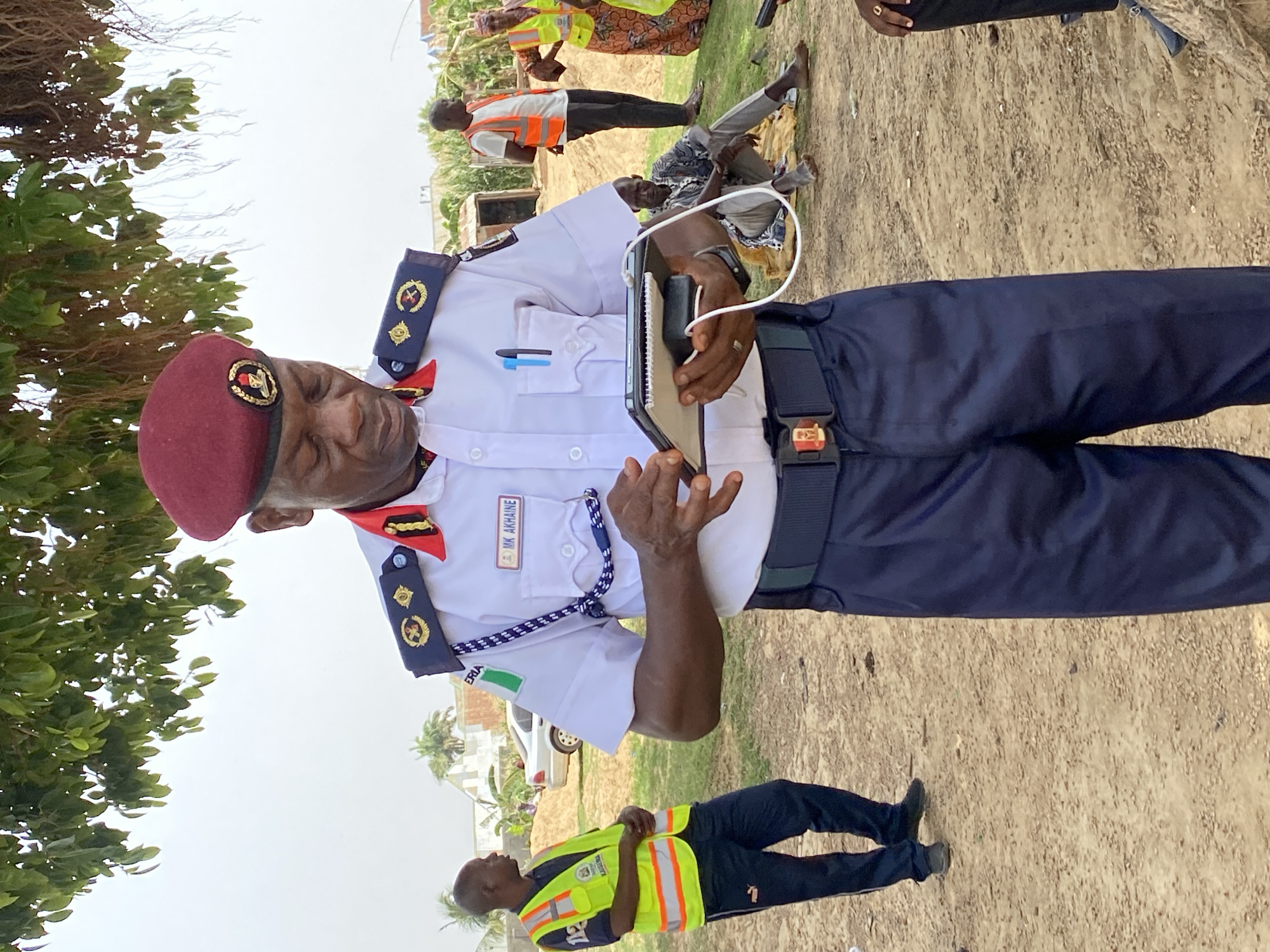 A man in a maroon beret and a white uniform is holding a cell phone. He is standing in a dirt lot with other people. Some of the other people are wearing safety vests. The photo was taken in Rigachikun, Nigeria.