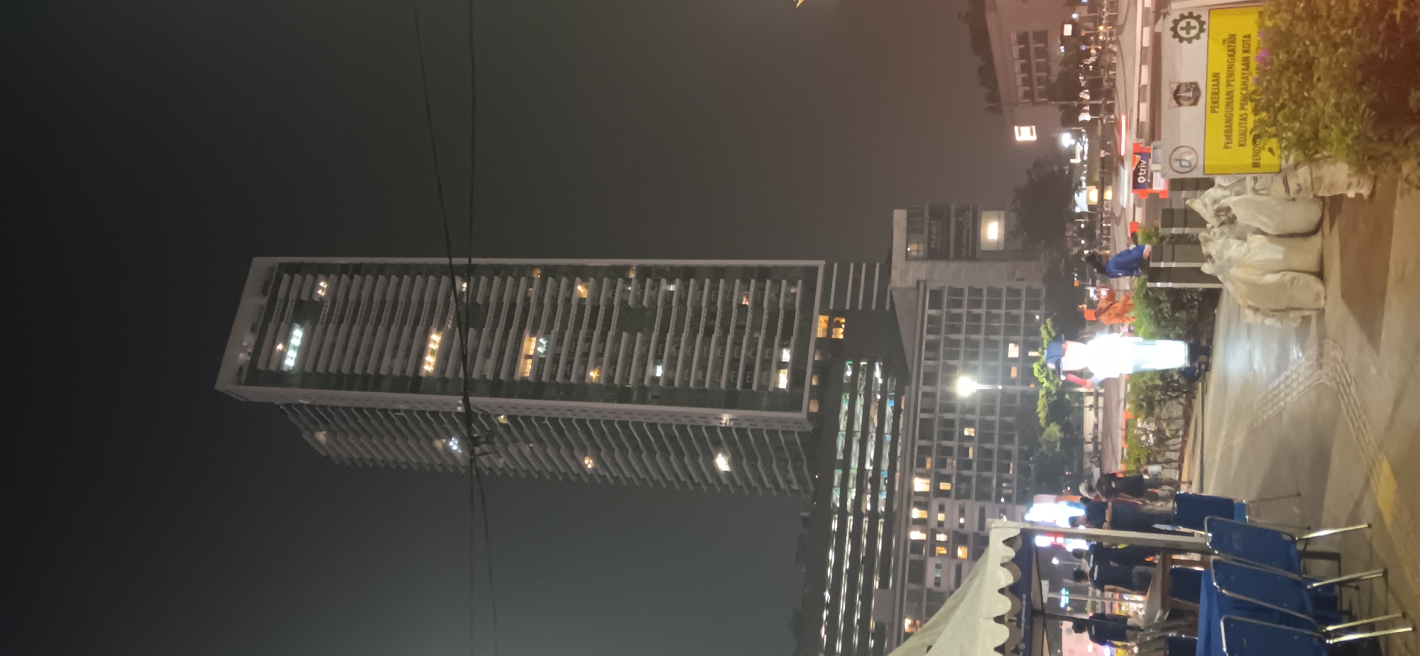 This is a nighttime, vertical-format, low-light photograph taken in Central Jakarta, Indonesia. The primary subject is a tall, modern skyscraper with a white facade and numerous floors, each featuring horizontal balconies and rectangular windows, many of which are illuminated. To the left of the skyscraper, a lower, wider building with a grid-like pattern of windows is visible. In the foreground, a white tent with a blue trim is partially visible on the left. Several people are gathered near the tent, some standing and conversing, others appearing to be working. Blue chairs and a table with a blue tablecloth are set up in front of the tent. A paved walkway with a tactile paving pattern runs horizontally across the lower part of the image, bordered by a small hedge. Several large, white sacks are piled on the ground to the right of the walkway. A yellow sign with black text, written in Indonesian, is propped up near the sacks, indicating construction or improvement work. Further in the background, streetlights, traffic signals, and the outlines of other buildings are visible, suggesting an urban environment with ongoing activity. The sky is dark and hazy.