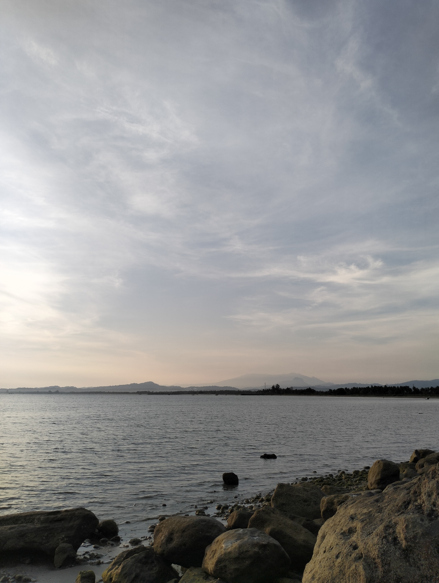 The image depicts a seascape in Lhokmanee, Indonesia, under a cloudy sky. The foreground features large, greyish-brown rocks, some partially submerged in the water. The water is calm with small ripples. In the midground, the shoreline is visible, marked by a thin strip of land with trees. The background reveals a distant mountain range, hazy and indistinct. No people are visible.