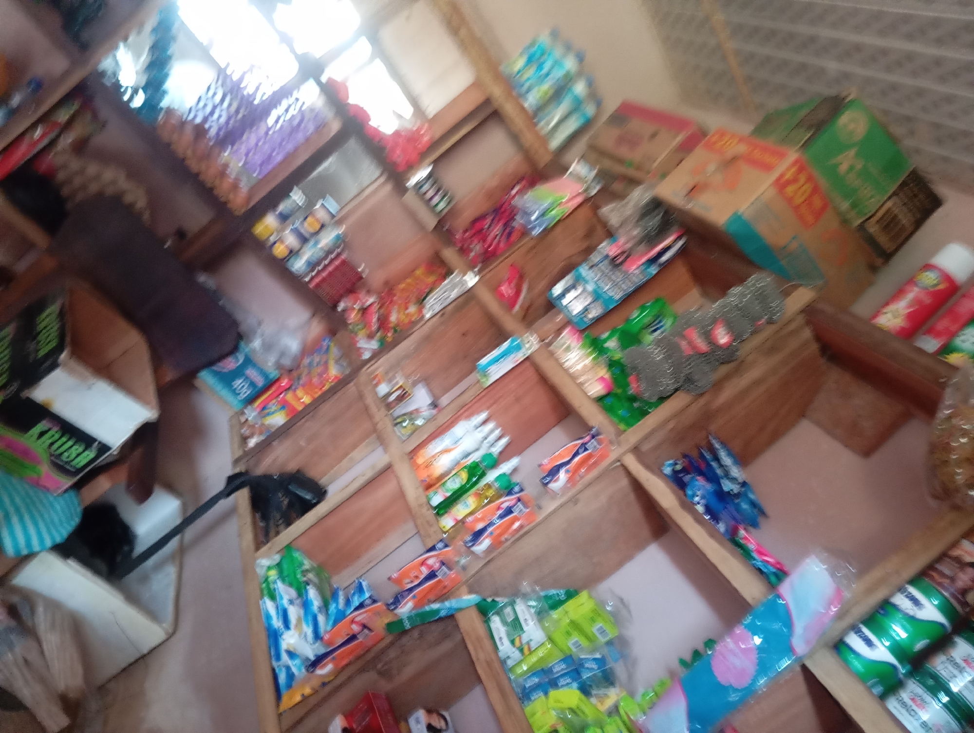 The image shows the interior of a small shop, likely in Jalingo, Nigeria, with wooden shelves stocked with various goods. The shelves are filled with a diverse range of products, including bottled beverages, boxed items, snacks in colorful packaging, and some cleaning or personal care products. Some of the visible items include bottles of Coca-Cola, bottles of water, and bags of snacks. The lighting suggests it could be daytime, with light coming from windows or an open doorway. The arrangement of the products suggests a retail environment where items are displayed for sale. The overall impression is of a typical local convenience store or small market.