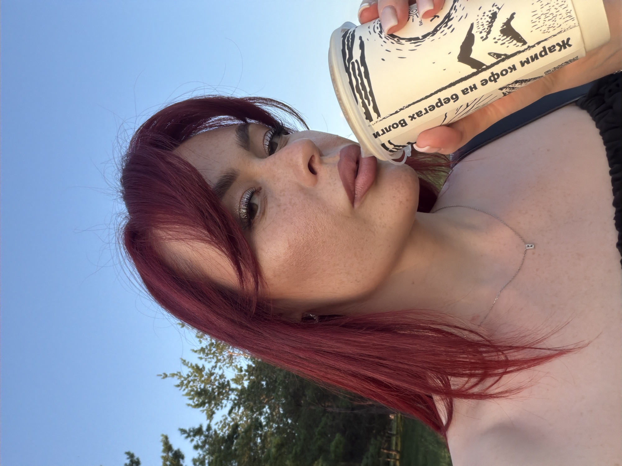 In the city of Tolyatti, Russia, a young woman with vibrant red hair is captured in a close-up selfie outdoors. She is holding a paper coffee cup with a black and white design in her right hand, bringing it to her lips as if taking a sip. Her left hand is visible near the cup, with a pale pink manicure. The woman has a light complexion with freckles across her face and wears makeup, including shimmery eyeshadow and lip gloss. A delicate silver necklace with a small rectangular pendant rests on her collarbone. She is looking towards the camera with a relaxed expression. The background consists of a clear blue sky and the tops of green trees, suggesting a park or a natural setting.