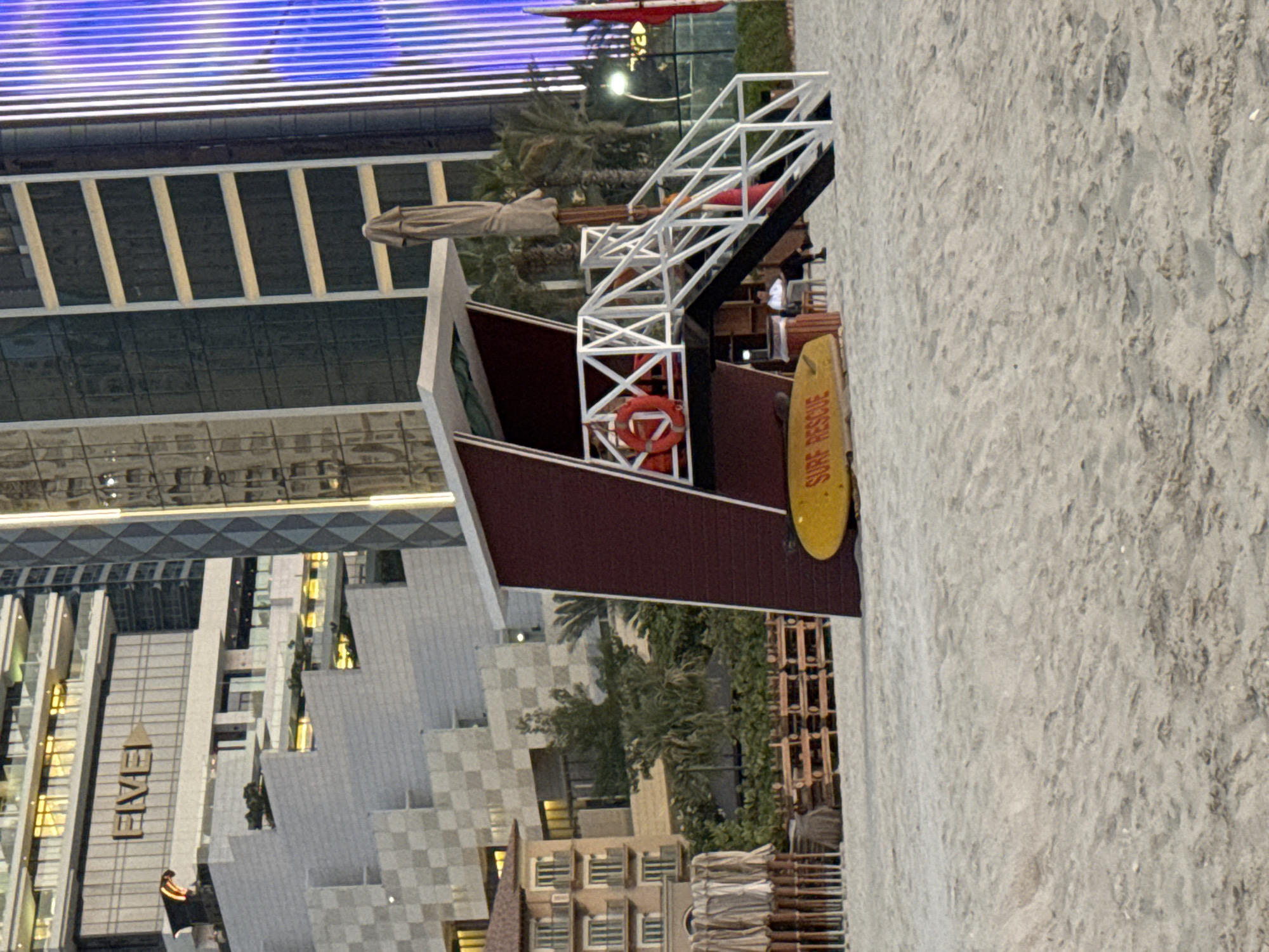 The image captures a beach scene in Dubai, United Arab Emirates, likely near the FIVE Palm Jumeirah hotel, as indicated by the visible "FIVE" signage on one of the buildings. In the foreground, a red and white lifeguard tower stands prominently on the sand. A yellow surf rescue board is placed in front of the tower. The tower has a white railing and a ladder leading up to a platform. A red life preserver is hanging on the railing. Behind the lifeguard tower, several rows of folded beach umbrellas are visible, suggesting a resort or hotel area. In the background, modern high-rise buildings with glass facades dominate the skyline, characteristic of Dubai's urban landscape. The sand appears fine and light-colored, with subtle footprints. A few palm trees and other greenery are interspersed between the buildings and the beach. The lighting suggests it is either early morning or late afternoon. Two individuals are seated near the base of the lifeguard tower, one wearing a white shirt and the other in dark clothing.
