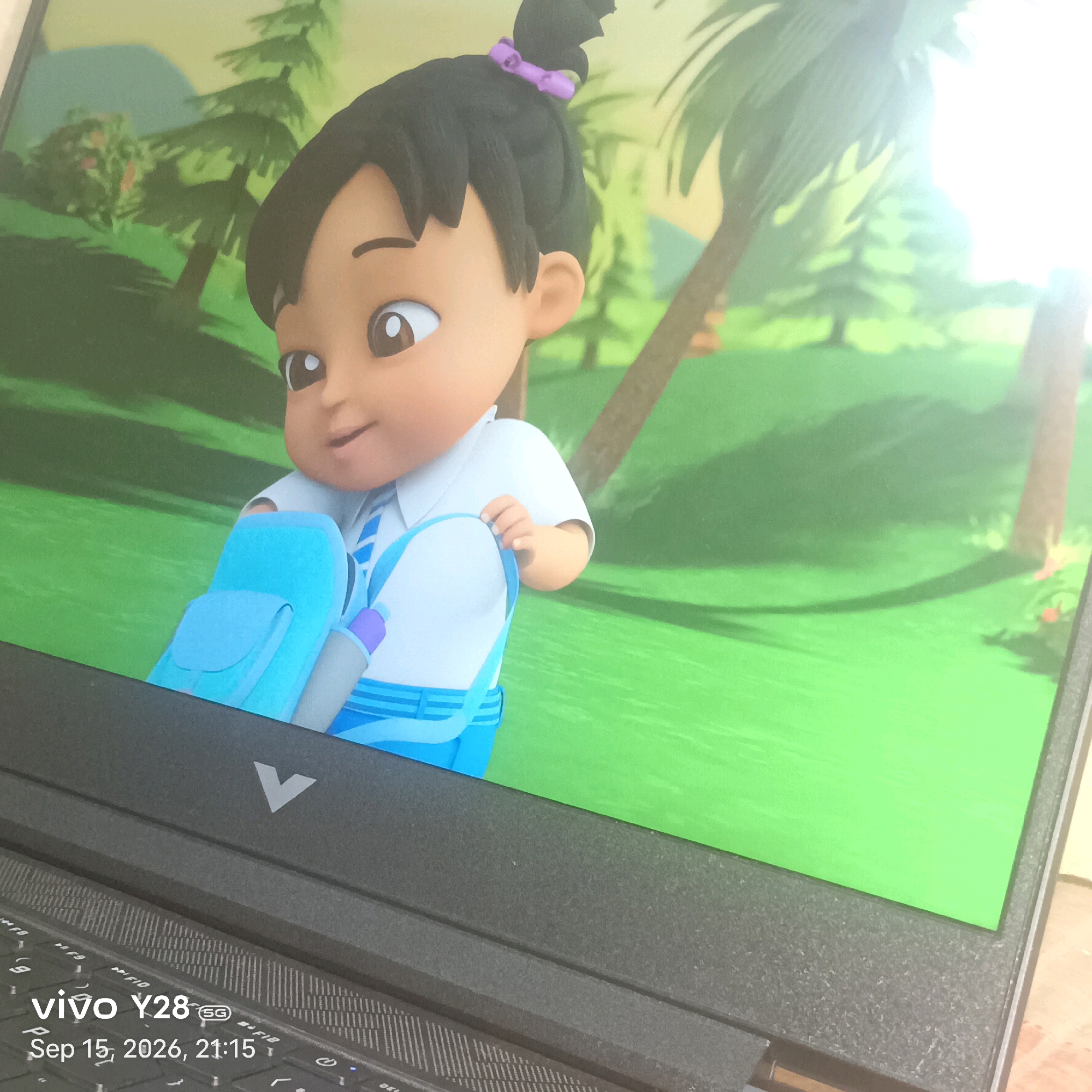 An animated girl wearing a school uniform, consisting of a white shirt and a blue and white striped tie and skirt, is depicted on a laptop screen. She has black hair tied up with a purple ribbon and is carrying a light blue backpack. The background on the screen shows a cartoon landscape with trees and a bright green grassy area. In the foreground, the keyboard of a laptop is visible, with a "vivo Y28 5G" logo and the date "Sep 15, 2026, 21:15" imprinted on it. The laptop's logo, a stylized 'V', is also visible. The image appears to be taken indoors, with the laptop placed on a textured surface. There is no discernible relation to Alirajpur, India.