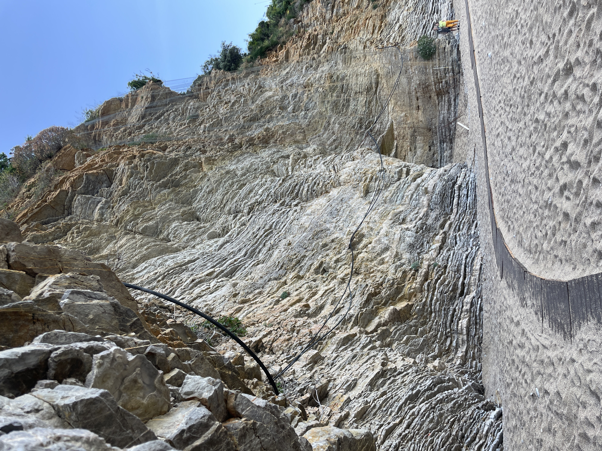 The image depicts a steep, rocky hillside in Sveti Stefan, Montenegro. The hillside is composed of light-colored, striated rock formations. A black pipe runs along the base of the hillside, partially obscured by large, gray rocks in the foreground. Above the primary rock face, there's a section with a net preventing rocks from falling. At the top of the hillside, some greenery is visible. To the right of the hillside, there's a portion of a concrete or stone structure. At the top of the concrete structure, a person wearing an orange shirt and light-colored pants is located on the edge. A series of cables is also visible connecting to the cliff.