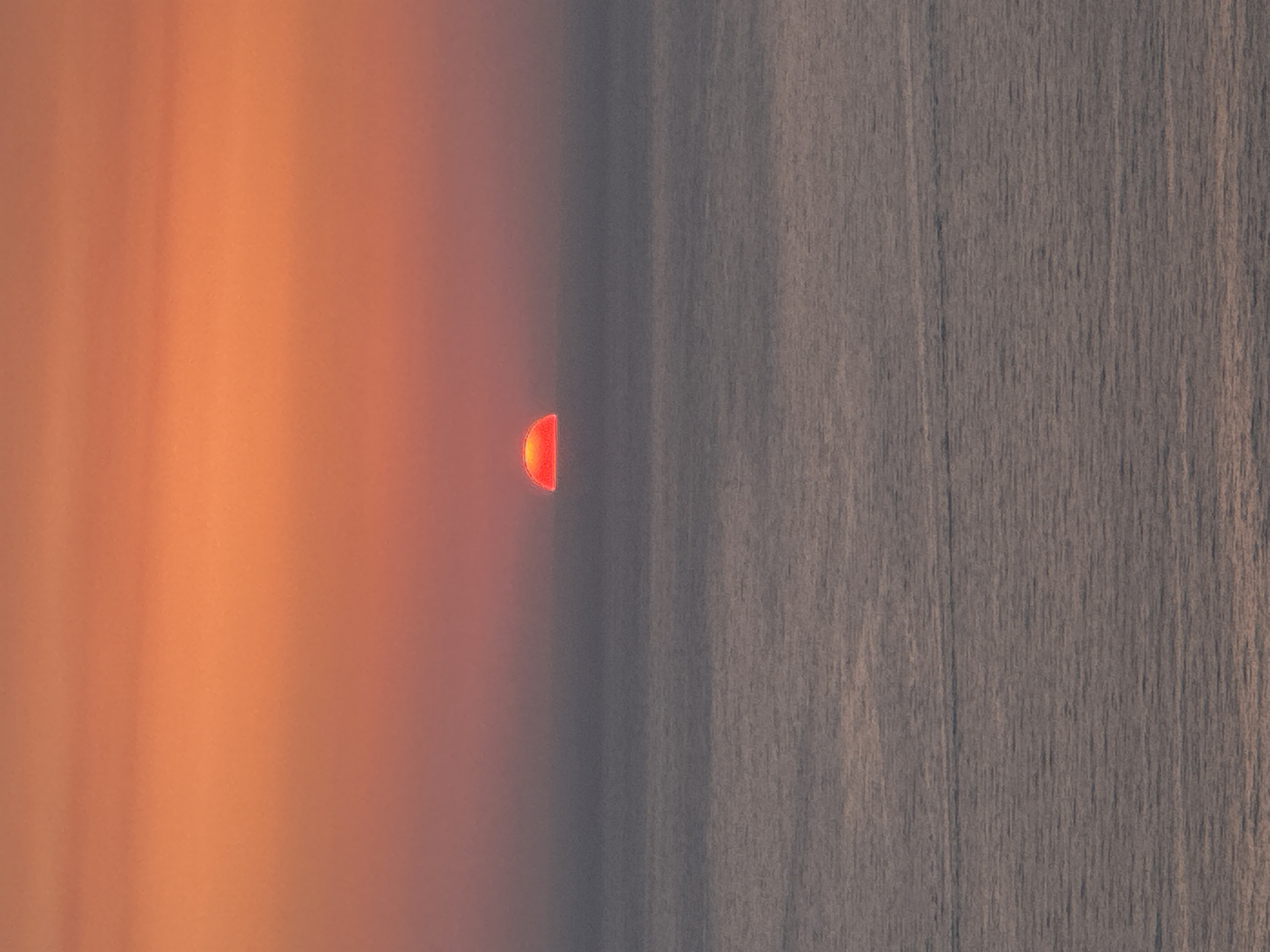 A vertical, medium shot captures a sunset over the Adriatic Sea. The sun, a vibrant orange-red orb, is half-submerged on the horizon, casting a fiery glow across the sky. The sky transitions from a bright orange at the top to a softer, hazy orange closer to the sea. The water below is a muted gray, with subtle horizontal lines indicating gentle waves. The image is devoid of any visible people, man-made structures, or identifiable landmarks, making it impossible to ascertain a specific location within Zagora, Montenegro, or any city. The scene is purely natural, focusing on the atmospheric spectacle of the setting sun.