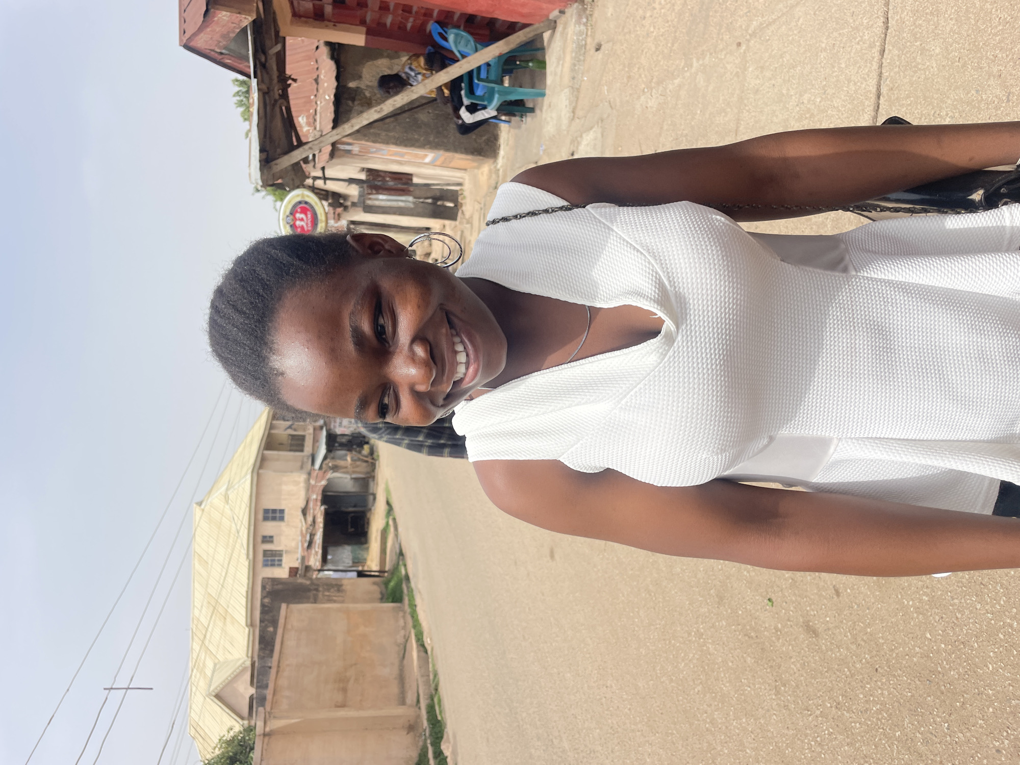 A young woman is standing on a paved street in Jos, Nigeria. She is wearing a white dress and silver hoop earrings, and she is smiling at the camera. The background includes buildings with corrugated metal roofs and a blue plastic chair, indicating a mixed residential and commercial area. Power lines are visible against the sky.
