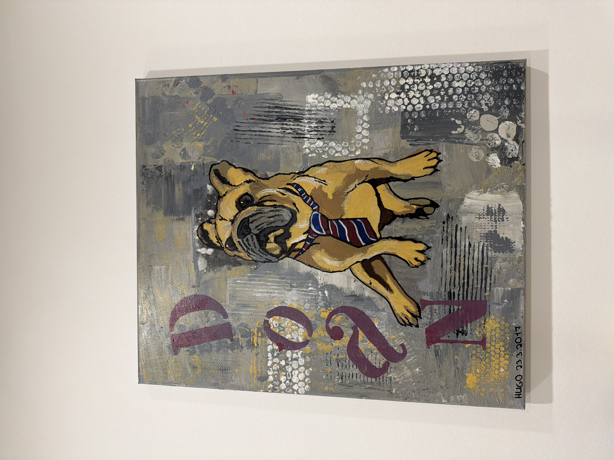 A rectangular canvas featuring an abstract painting is mounted on a white wall. The painting depicts a tan dog wearing a blue and red striped tie, rendered in a cartoonish style. Below the dog are stylized, stenciled letters "DOG." The background consists of gray and white paint with dotted patterns and black hash marks. The painting is signed "HUGO 23.3.2017" on the right edge. The image does not contain information to determine the city of Pétange, Luxembourg is located.