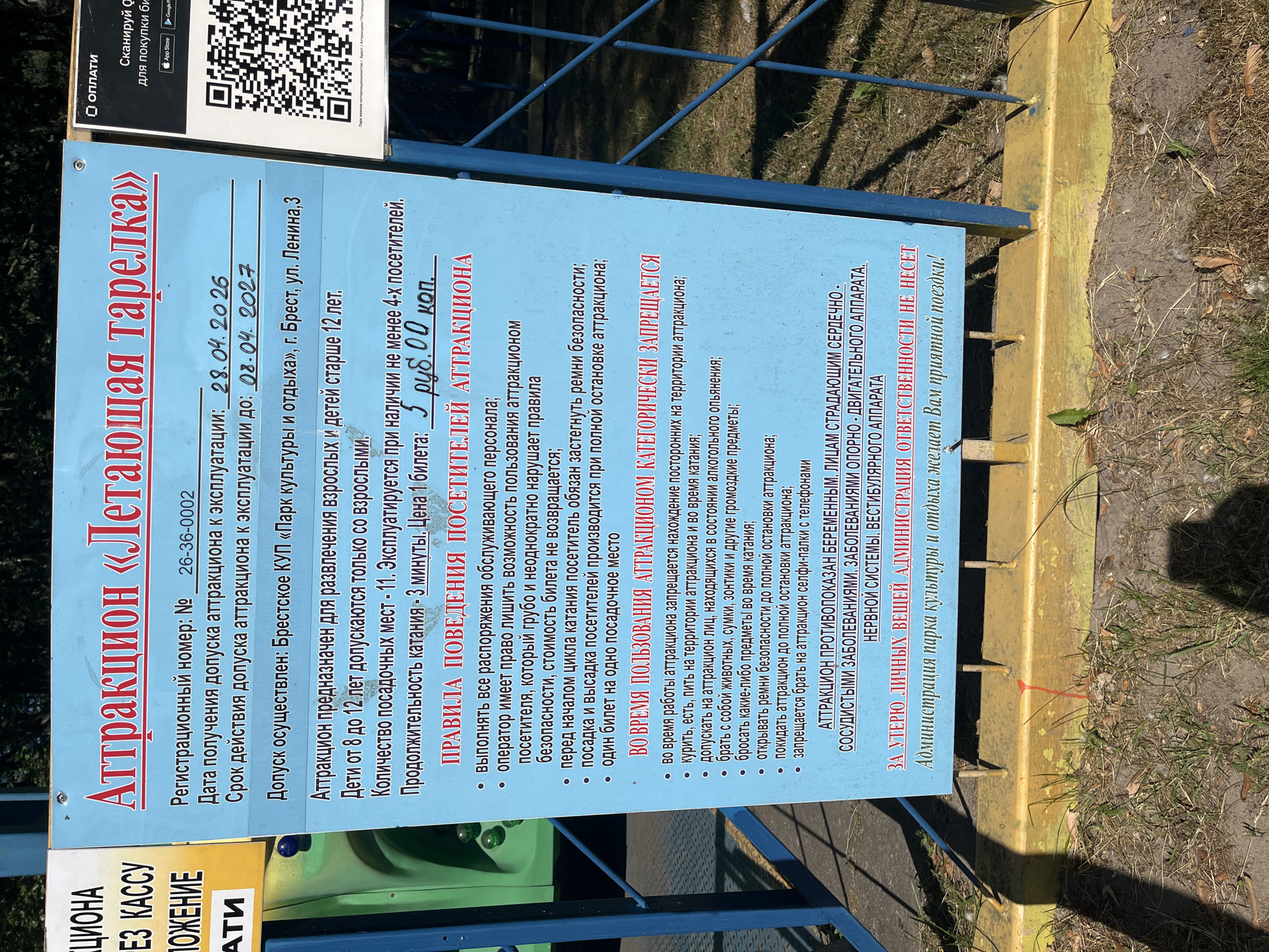 The image shows a blue sign with white and red lettering, likely from an amusement park. The sign is mounted on metal poles and appears to be informational. The text on the sign is in Russian and describes an attraction called "Flying Saucer." It details rules and regulations for visitors, including age restrictions, safety guidelines, and pricing. The sign also has a QR code for payment. The ground around the sign is covered in grass and dirt. The overall scene suggests an outdoor setting at a recreational facility.
