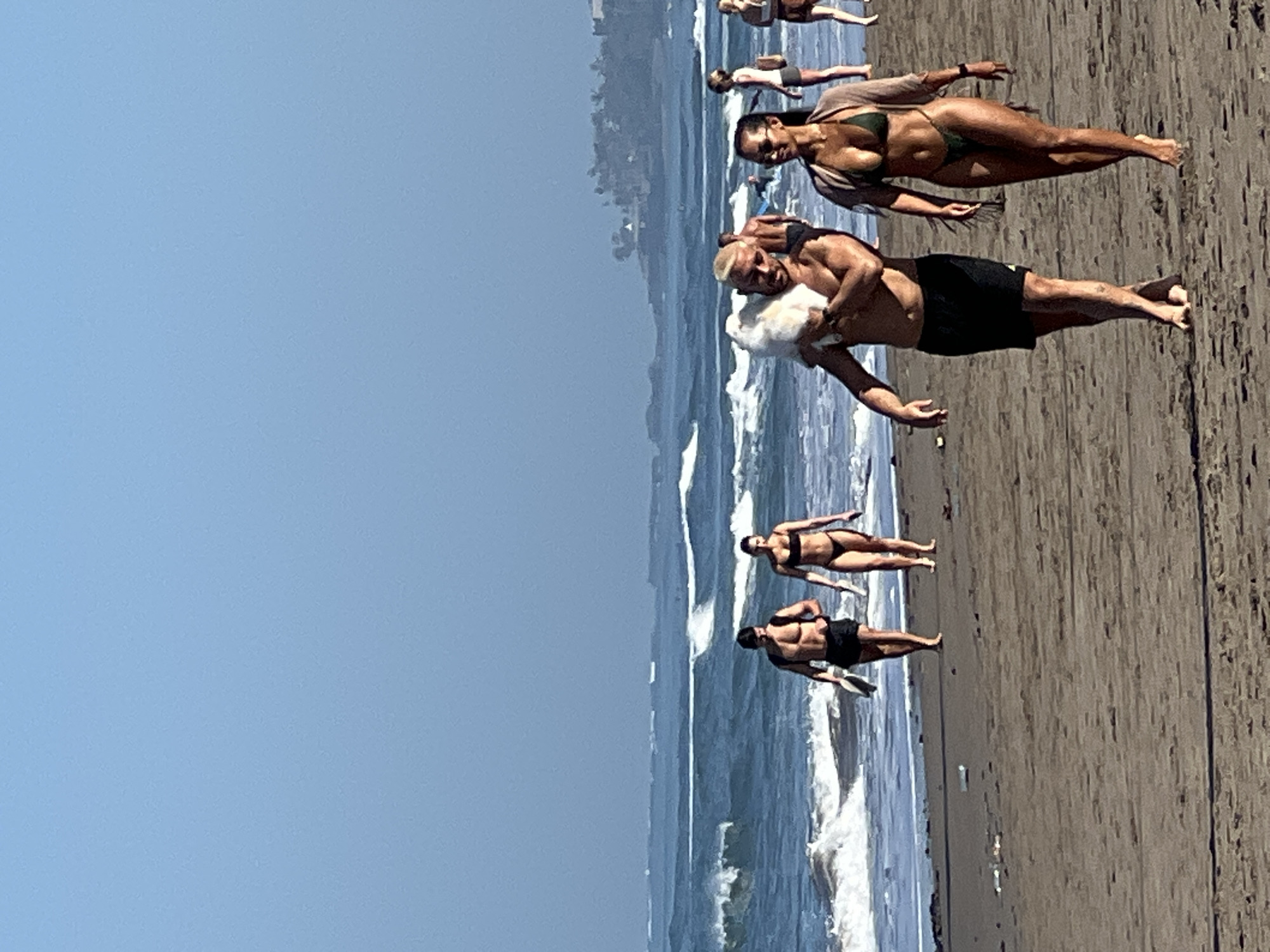 In North Kuta, Indonesia, on a sandy beach, a man with blond hair and muscular physique walks towards the viewer, carrying a fluffy white animal over his shoulder. He wears black swim shorts. Beside him, a woman with dark hair and sunglasses walks to his right, wearing a dark green bikini and a sheer, fringed wrap. Behind them, other individuals are visible on the beach and in the ocean. Several people in swimsuits are walking along the shore in the mid-ground, while the background features the ocean with waves breaking and a distant coastline with vegetation and buildings. The sky is a clear, pale blue. The sand is light brown, with some debris scattered on its surface.