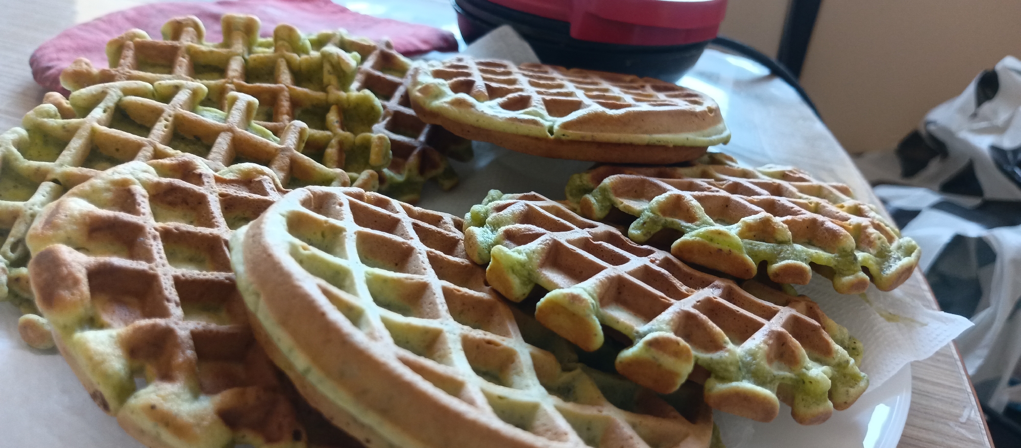 The image shows a stack of green-tinted waffles on a white plate. Some waffles are overlapping, and they have a golden-brown color on the outside with a greenish hue on the inside. A waffle maker with a red handle is partially visible in the background. A pink cloth is also in the background, slightly blurred. The scene appears to be indoors, possibly in a kitchen setting.