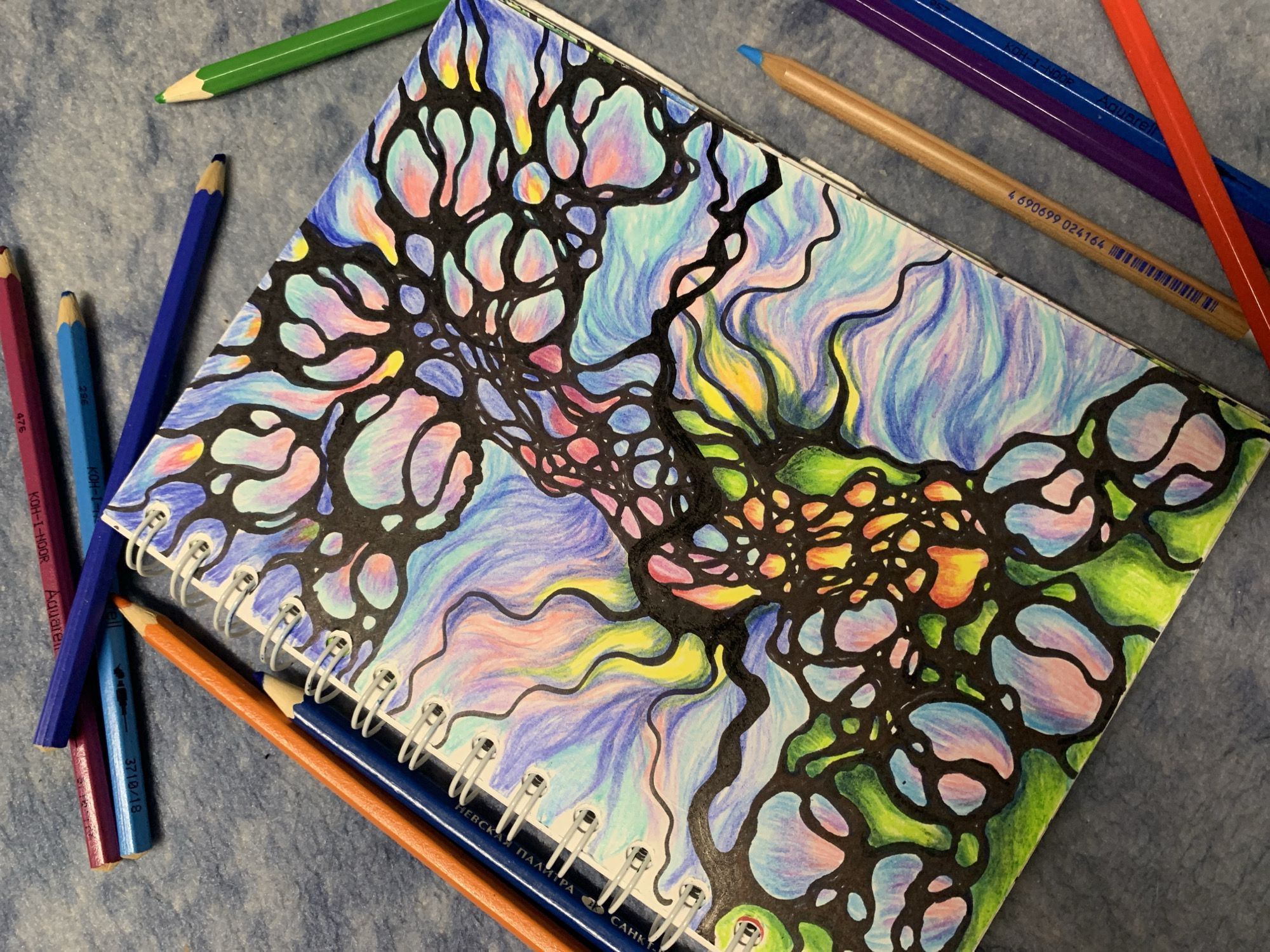 This media file captures a close-up, overhead view of a sketchbook open to a vibrant, abstract drawing. The drawing features a black, intricate, branching pattern filled with swirling lines of color, predominantly blues, purples, pinks, yellows, and greens. The style suggests a neurographic art technique, creating a sense of organic movement and energy.
Scattered around the sketchbook are several colored pencils, including a green pencil at the top left, a blue pencil to the left of the drawing, a purple pencil and a red pencil towards the top right, and a tan pencil with a barcode and numbers "46906990241648" printed on it, also near the top right. A blue pencil and an orange pencil lie near the bottom edge of the sketchbook. The sketchbook itself is spiral-bound with white rings and has text visible on its blue cover at the bottom: "НЕВСКАЯ ПАЛИТРА САНКТ-".
The lighting suggests an indoor setting, likely a flat surface like a table or desk, given the blue-grey textured background that resembles a fabric or mat. There are no people present. The overall impression is one of creative activity, with the drawing and pencils laid out as if recently used or about to be used for artistic expression. The mood conveyed is one of artistic focus and colorful imagination.