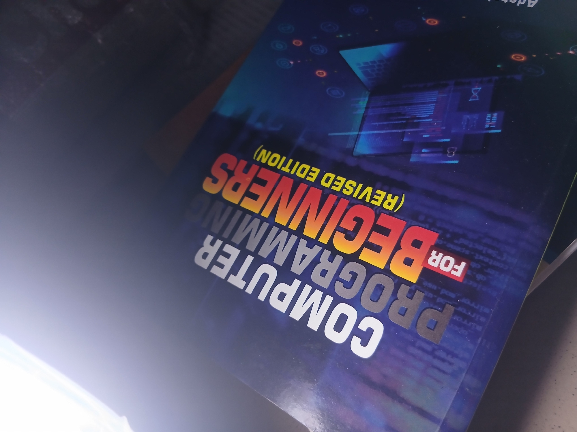 A book titled "COMPUTER PROGRAMMING FOR BEGINNERS (REVISED EDITION)" is angled in a portrait orientation, with the top left corner illuminated by bright light, suggesting it is a photograph of the book's cover. The cover features a dark blue background with abstract digital patterns and a laptop illustration. The title is prominently displayed in large, bold letters. The words "COMPUTER PROGRAMMING" are in white and gray, stacked vertically. "FOR" is in a red box. "BEGINNERS" is in large, bold, orange-red letters, angled diagonally across the cover. Below "BEGINNERS," in yellow text, is "(REVISED EDITION)". The lighting creates a glare that partially obscures the upper left portion of the image. The book is resting on a dark surface. There are no people or animals visible. The setting is indoors, and the specific location within a city is unknown.