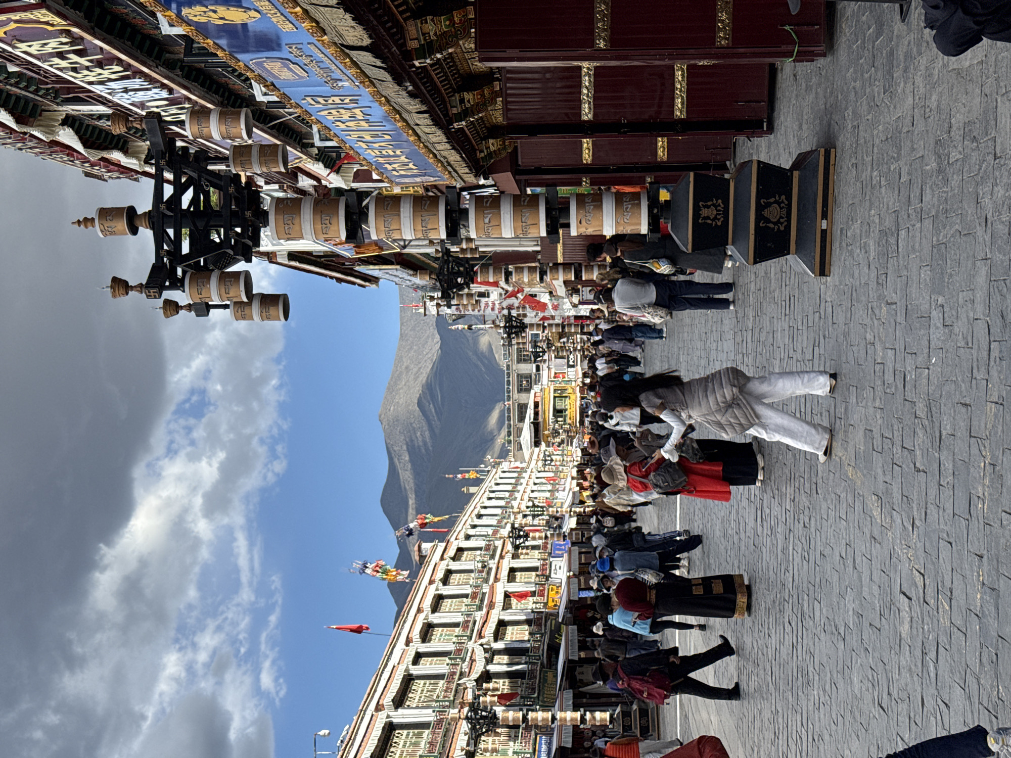 The image depicts a vibrant street scene in a Tibetan cultural area, likely within Chengguan District, China. The perspective is street-level, looking down a paved thoroughfare flanked by traditional multi-story buildings and a mountainous backdrop.
In the left foreground, a prominent structure features numerous large, cylindrical prayer wheels. These prayer wheels are light brown with visible Tibetan script inscribed on them, and some smaller ones are mounted on a central black frame decorated with golden swastika-like symbols (a Buddhist emblem). Above these, the buildings showcase intricate architecture with blue banners displaying Chinese characters and ornate gold and red decorative elements.
The street itself is paved with grey interlocking stones and is bustling with numerous pedestrians. People are seen walking, many appearing to be engaged in a circumambulation or "kora," possibly turning the prayer wheels as they move. Their attire is a mix of traditional robes and modern clothing, with some individuals carrying bags or backpacks.
In the mid-ground and background, a large, bare mountain range rises prominently under a dynamic sky. The sky transitions from clear blue towards the horizon to patches of white and darker grey clouds directly overhead. Colorful prayer flags and red flags are visible fluttering from the rooftops of the buildings, adding to the cultural atmosphere.
On the right side of the street, more traditional buildings with dark red facades and golden accents are present. Near these structures, black decorative boxes adorned with golden symbols are placed on the ground. The scene conveys a sense of bustling activity within a culturally rich and picturesque setting, suggesting a place of spiritual significance and daily life.
