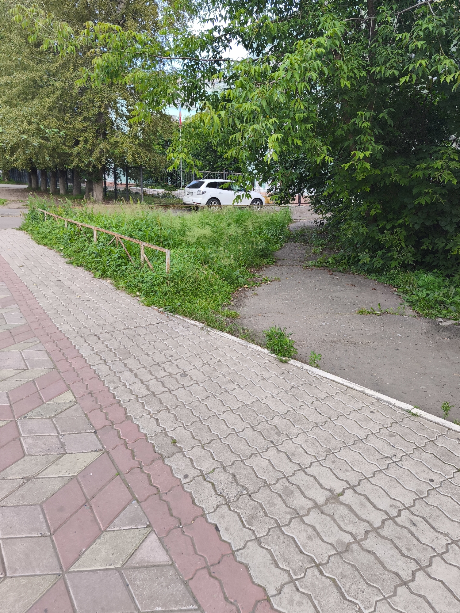 The image depicts a paved walkway in Komsomolsk-on-Amur, Russia. The walkway is constructed with interlocking concrete pavers in a grey geometric pattern, bordered on one side by a section of pavers in a pinkish-red and grey diamond pattern. To the left of the walkway, a weathered metal fence with a reddish-brown patina runs along a strip of overgrown green grass and weeds. Further beyond the grass, a row of trees with dense green foliage is visible, and behind them, a white SUV is parked. A grey asphalt path branches off to the right of the walkway, leading into an area with more lush green vegetation and trees. The overall scene suggests an outdoor urban environment, possibly a park or residential area, with a focus on the walkway and surrounding greenery.