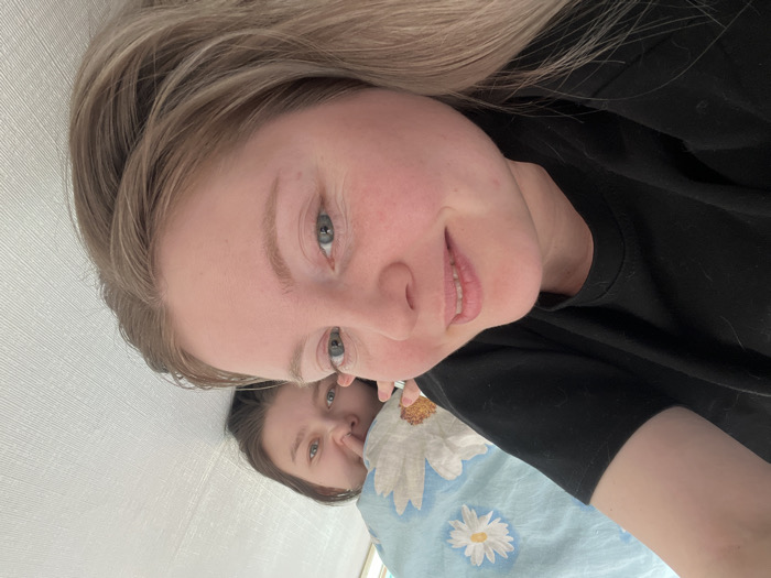 The image displays two young adult females positioned horizontally in an indoor residential setting, understood to be in Chaikovsky, Russia. The perspective indicates a close-up selfie taken from a prone position.
In the foreground, a female with blonde hair, fair skin, and light-colored eyes is partially visible from the upper torso, wearing a black t-shirt. She exhibits a slight smile and is looking directly at the camera.
Behind and to the left of the foreground subject, a second female is partially visible. She has darker hair, fair skin, and light-colored eyes, also directing her gaze towards the camera. This individual is partially covered by a light blue duvet patterned with white daisies, consistent with lying on a bed.
The background features a light-colored, subtly textured wall positioned to the right of the foreground subject and behind the background subject. The environment is illuminated by ambient, natural light.