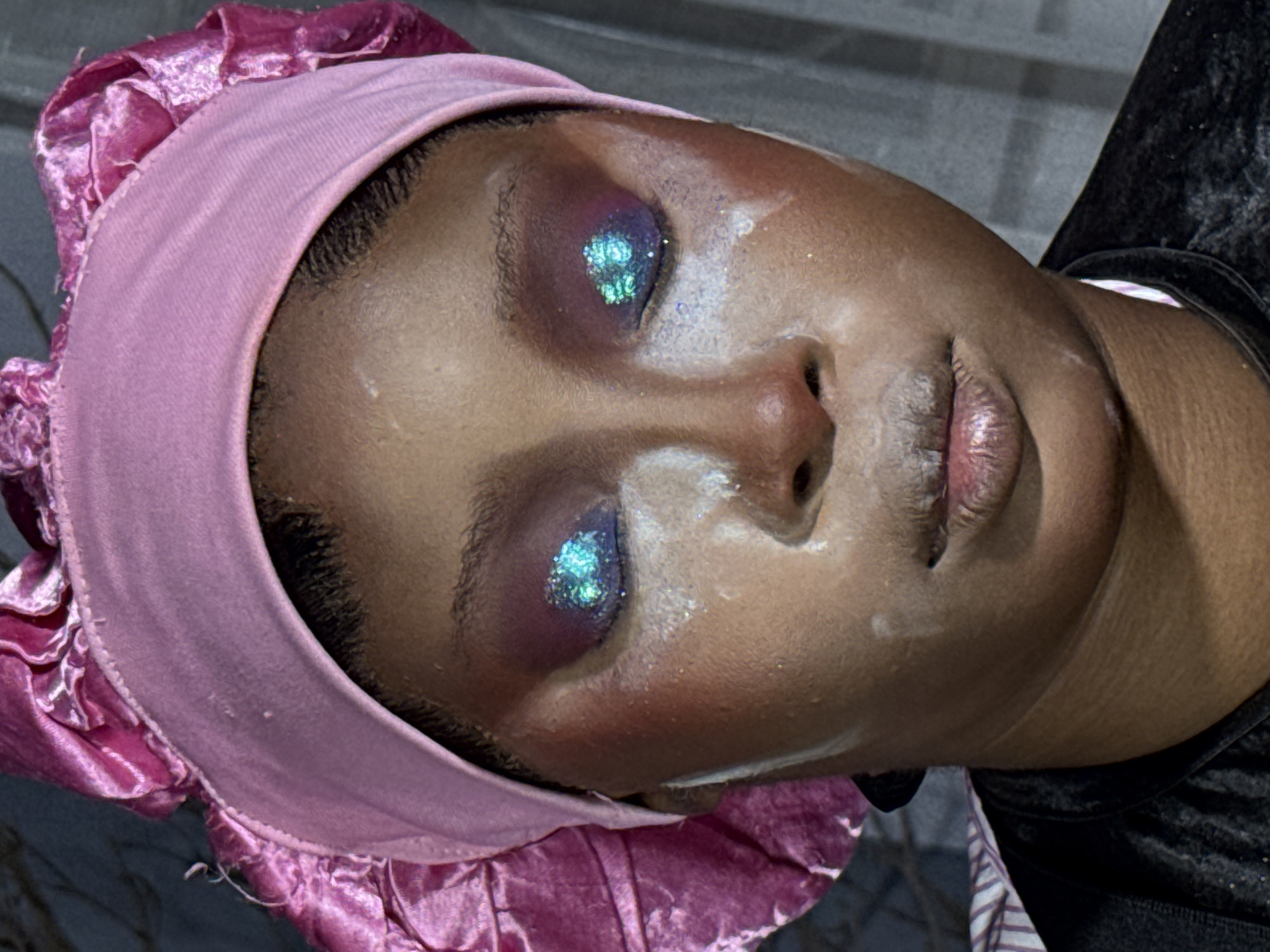 The image is a close-up, head-and-shoulders portrait of a Black person wearing a pink satin headwrap. The person has dramatic makeup on their eyes, featuring vibrant blue glitter on the eyelids and purple eyeshadow. White powder is applied liberally to the face, particularly around the eyes and nose, creating a stark contrast with the person's skin tone. The person's lips are slightly parted and appear to have a glossy texture with some of the white powder visible. The background is dark and out of focus, with vertical lines suggesting fabric or a curtain. The setting is likely indoors, given the controlled lighting. There is no discernible indication of the city of Gura Top, Nigeria.