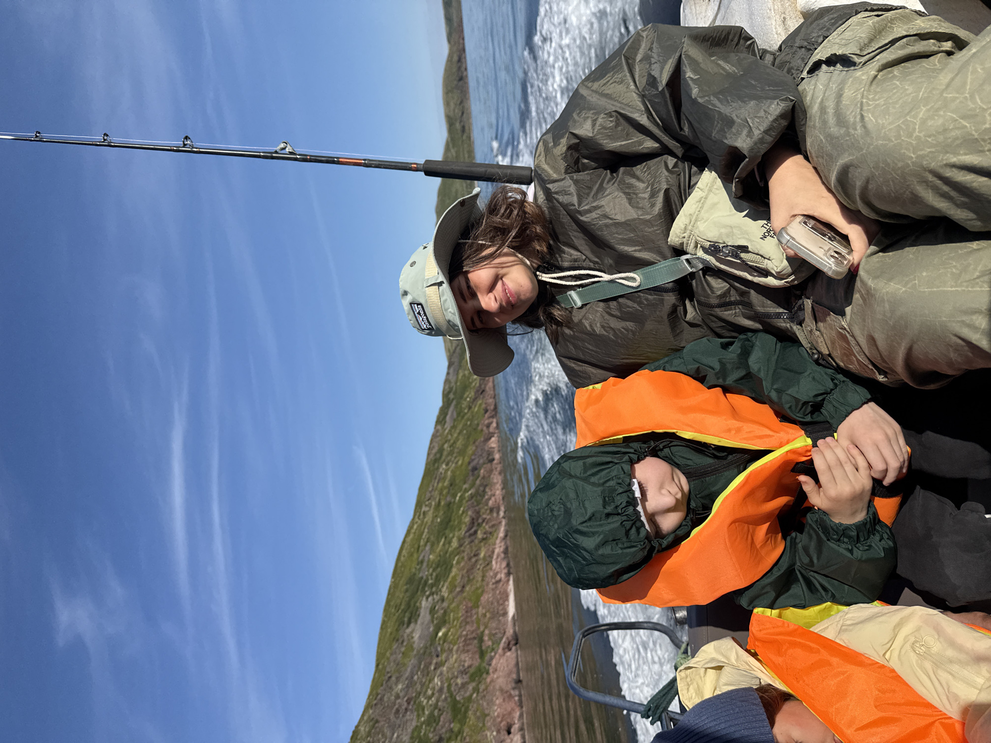 Two individuals are seated in a motorboat moving through the water, with a fishing rod extending upwards from the boat. The person in the foreground on the right is a young woman wearing a wide-brimmed hat, a dark green jacket, and a light-colored bag slung across her chest. She is holding a small silver device in her right hand. To her left, a child is wearing a dark green hooded jacket and an orange life vest. The child appears to be looking down, with their hands clasped. In the background, a rugged, green and reddish-brown rocky shoreline is visible under a bright blue sky with thin, wispy clouds. The boat is creating a wake as it travels through the water. The scene is consistent with the coastal landscape of Teriberka, Russia, a location known for its dramatic scenery and fishing opportunities.