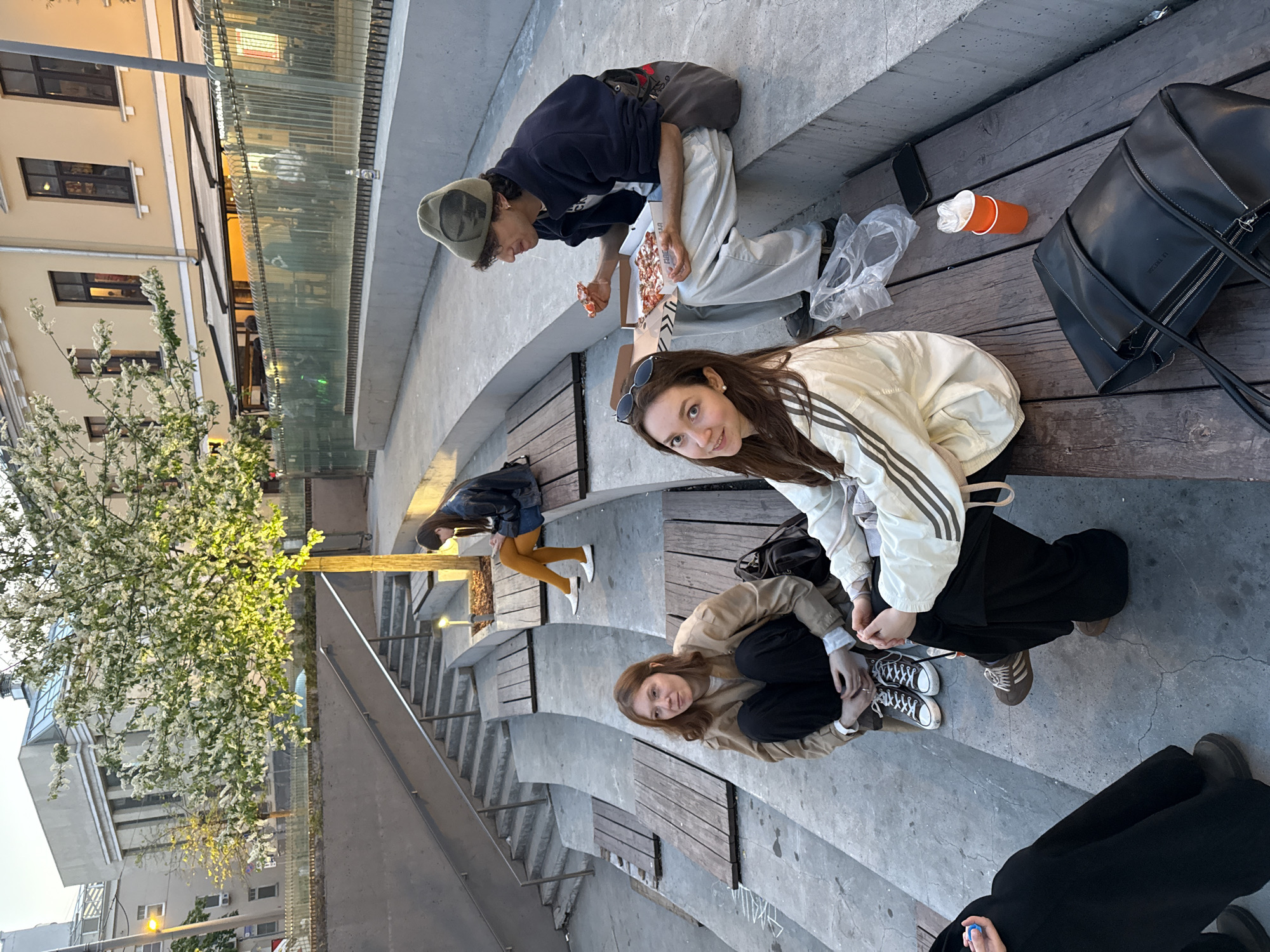 The image shows a group of people gathered in a public space, likely a tiered seating area or amphitheater-style structure. The location is the Zaryadye Park in Moscow, Russia.
Several individuals are sitting on wooden benches integrated into the concrete structure. Some are eating pizza, while others are interacting with each other. The clothing worn by the people suggests a casual, modern style. Various objects are scattered around, including a pizza box, plastic bags, cups, and a black bag.
The environment includes trees in bloom. In the background, there are buildings and structures, including a glass barrier. The overall atmosphere is that of a relaxed, informal gathering in an urban park setting.