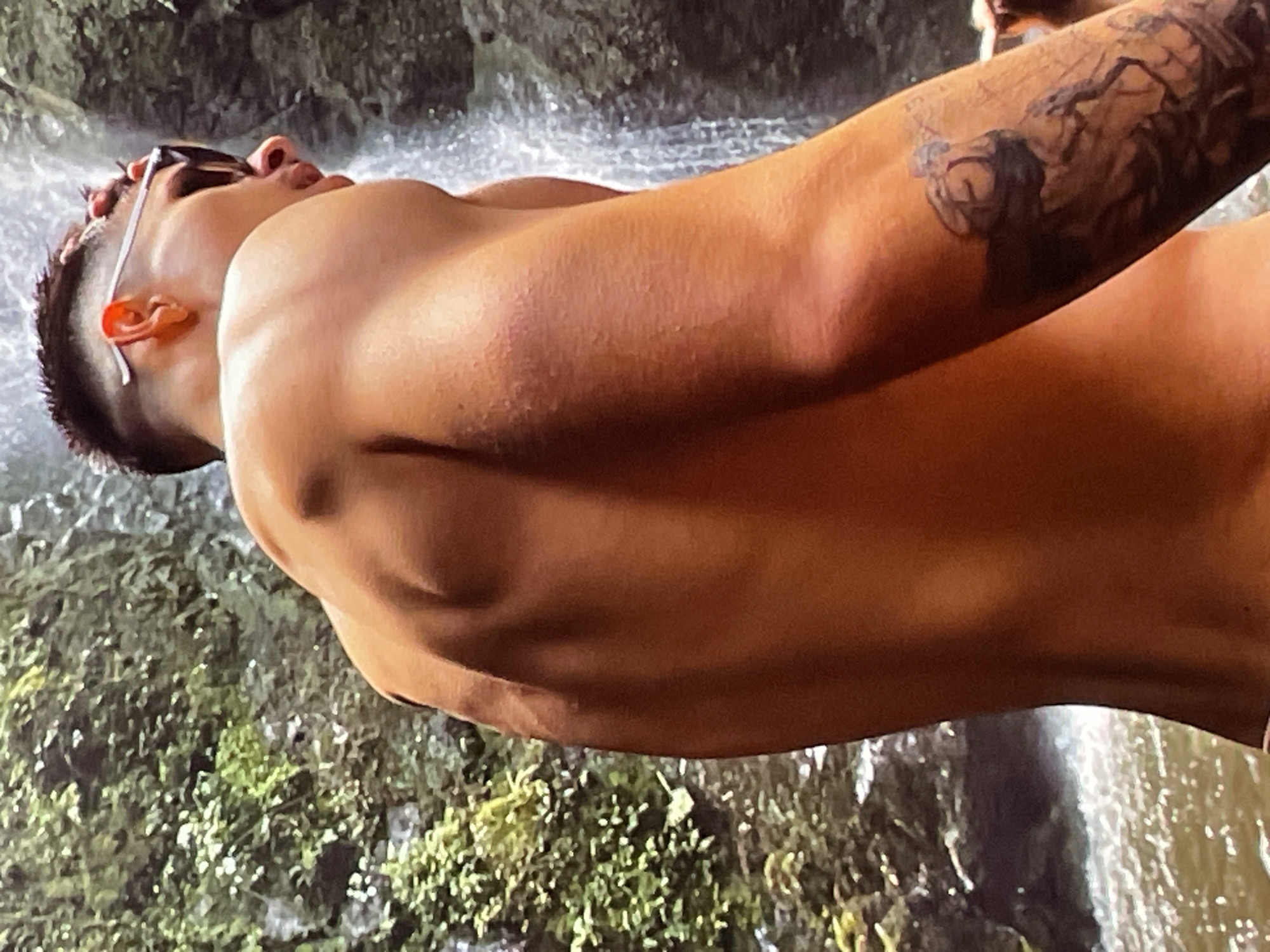 A man with short, dark hair and sunglasses resting on his forehead is standing in front of a waterfall. He has a tattoo on his arm and appears to be shirtless. The waterfall is surrounded by lush green vegetation. The location is likely near Harmanli, Bulgaria, given the visual cues of the natural landscape.