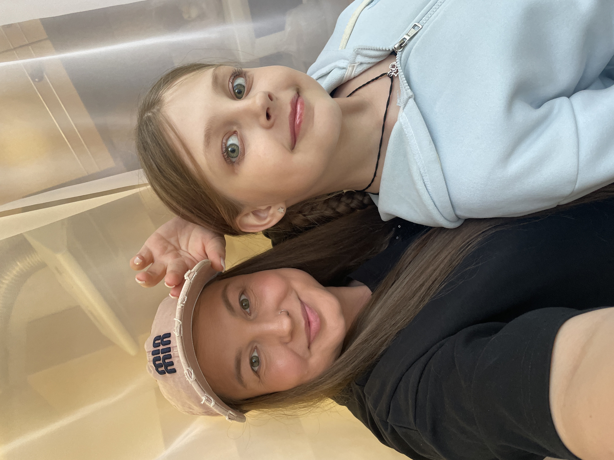Two females are in the picture. The female in the top center is a young girl with blonde hair and light blue sweatshirt and a black necklace. The female at the bottom is a younger women wearing a cap. The exact location in the city of Izhevsk, Russia, is not evident from the image. The background is plain and does not offer any clues to specific landmarks or locations within Izhevsk.
