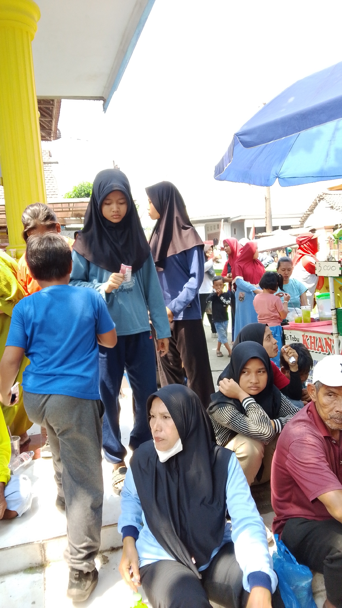 In Mancar, Indonesia, a bright outdoor scene unfolds with a gathering of people, likely at a local market or event. A prominent yellow pillar stands to the left, providing shade from the sun, which casts stark shadows. A blue tarped umbrella is visible in the upper right, suggesting a stall or vendor area.

The focus is on a group of individuals, many wearing hijabs. In the foreground, a woman in a black hijab and light blue long-sleeved shirt sits, looking to her left with a neutral expression. Beside her, a man in a maroon polo shirt also sits, looking forward. To the left of the woman, a young boy in a blue t-shirt and grey pants stands, looking away from the camera.

Further back, several young girls and boys are interacting. Two girls in black hijabs stand side-by-side. One, in a blue long-sleeved shirt, holds a small red item, possibly money. Another girl, in a darker blue top, stands next to her. Other individuals, including a woman in a red hijab and a woman in a light blue shirt behind a counter, are also visible. The counter appears to be a food or drink stall, with some containers and signage. The ground is paved and appears slightly dirty. The overall atmosphere is one of casual outdoor activity.