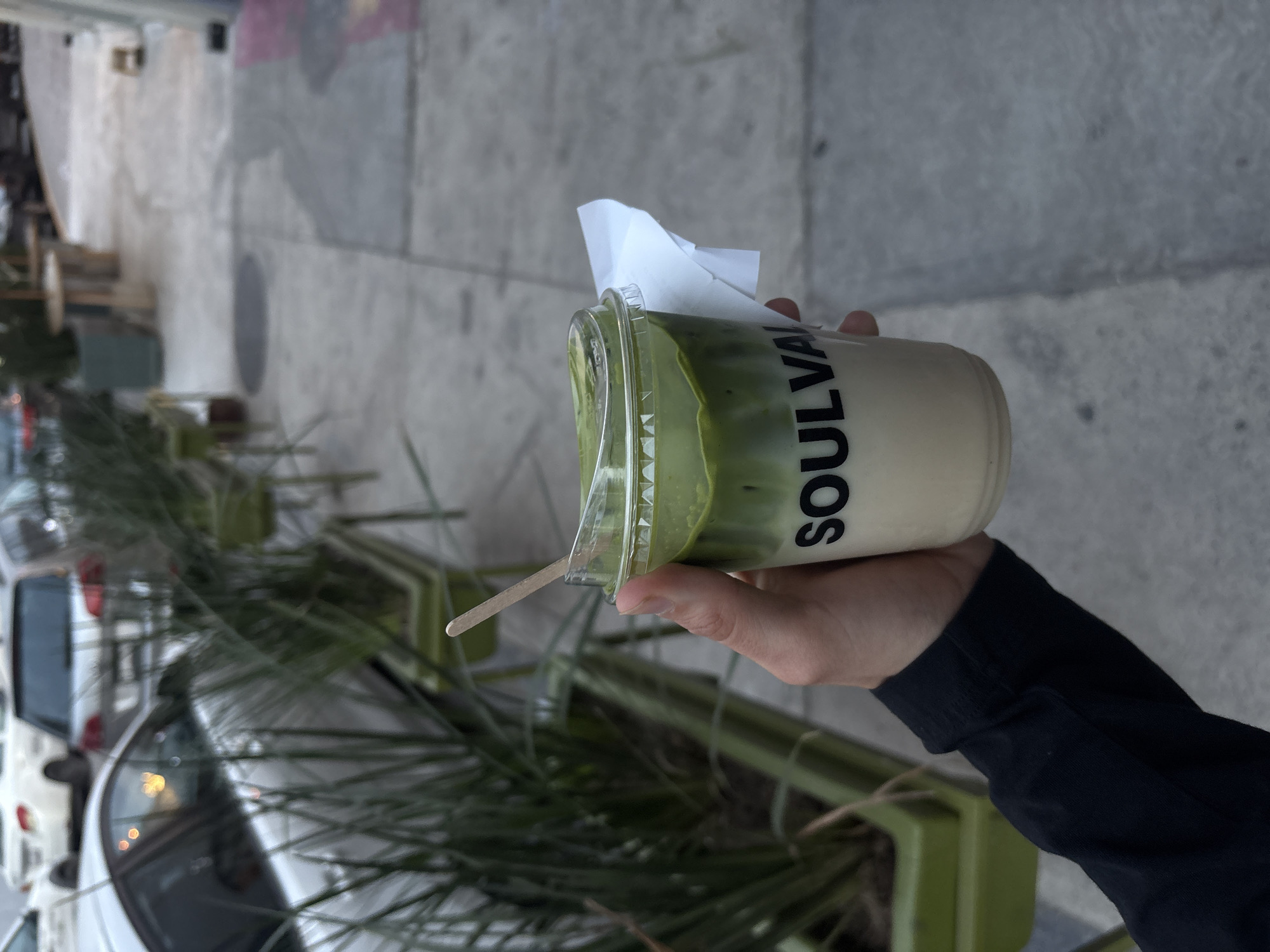 A person holds a plastic cup with a green and white beverage, bearing the name "SOULVA" in large, bold letters. The cup also contains a wooden stirring stick and a white paper napkin. The person's hand and a portion of a dark-sleeved arm are visible. The setting appears to be outdoors on a city sidewalk or street, as suggested by the concrete pavement, a glimpse of parked cars, and greenery in planters along the side of the street. It is possible this is the city of Puebla, Mexico.