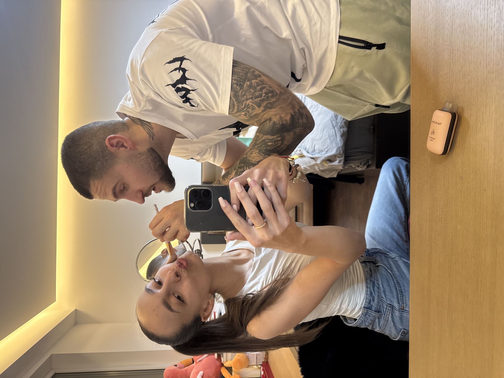 The image captures two young adults in a modern residential room in Sofia, Bulgaria, likely a bedroom or dressing area, with warm LED strip lighting illuminating a white ceiling. A female, seated on the left, is taking a mirror selfie with a dark-colored smartphone held in her left hand. She has long dark hair in a ponytail, wears a white sleeveless top and light blue denim jeans, and has a thin gold ring on her left ring finger. Her nails are manicured with light pink polish. She holds a small round mirror in her right hand, reflecting a portion of a male figure. The male, standing to her right, is leaning over and applying makeup to her face with a light-colored brush. He has short dark hair, a beard, a neck tattoo, and extensive sleeve tattoos with forest-like motifs. He wears a white short-sleeved t-shirt with black abstract designs on the shoulders and light green cargo shorts, with multiple bracelets on his left wrist. In the foreground, on a light wooden surface, a pink and black ELF BAR vape device is visible. In the background, to the left of the female, a pink flamingo plush toy and an orange object are visible, with a bed and duvet further back.