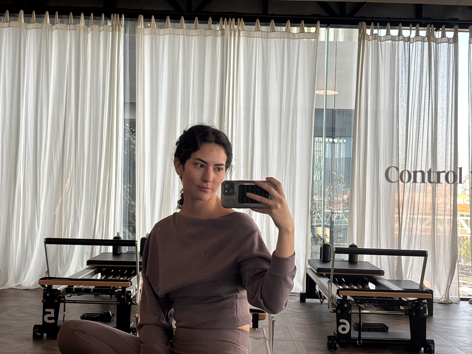 A woman is taking a selfie in a Pilates studio. She has dark curly hair and is wearing a matching mauve-colored crop top and leggings. She is holding her phone in her right hand, partially obscuring her face. Behind her are two Pilates reformer machines. The studio has light wood floors and large windows covered with light-colored curtains. The reflection in the window suggests a city view with some buildings and greenery visible. The text "Control" is visible on the right side of the image, possibly part of a logo or sign in the studio. The location is not San Bernardino Tlaxcalancingo, Mexico.