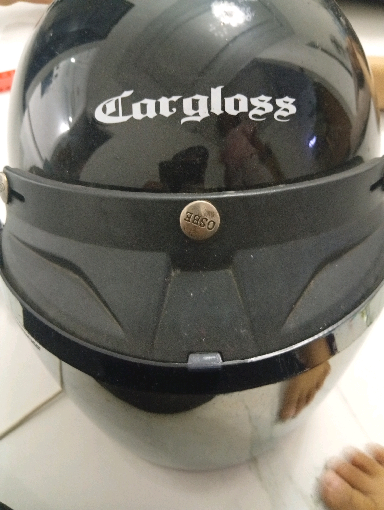 This is a close-up, head-on shot of a black motorcycle helmet, likely taken indoors on a white surface. The helmet features a white logo in a gothic font that reads "largloss" on the front, above a black brim. There are two circular metallic studs on either side of the brim, each with the letters "OSBE" embossed on them. The helmet is reflective, showing a blurred reflection of what appears to be a person's feet and hands, as well as a light source. There are some minor scuff marks and dust visible on the helmet's surface. The lighting suggests an indoor environment, possibly a home or workshop, rather than an outdoor urban setting. The image does not provide any visual cues or context to place it specifically within the city of Samarinda, Indonesia.