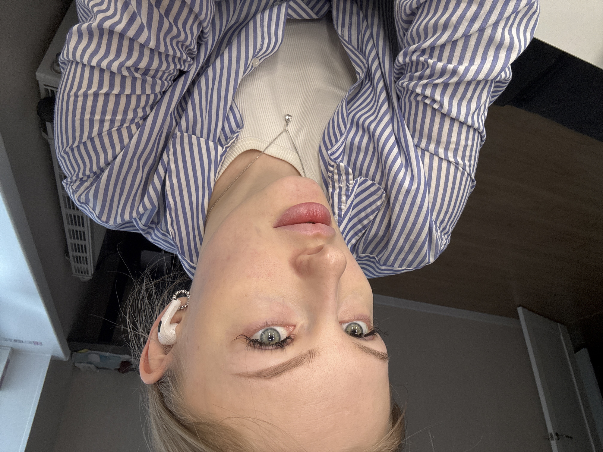 A woman is viewed from a close, upward angle, wearing a blue and white striped, unbuttoned shirt over a white ribbed tank top with a delicate silver necklace. She has blonde hair and is wearing a white wireless earbud in her left ear adorned with a jeweled ring. The background consists of an indistinct interior with light-colored walls and what appears to be furniture. There is no indication of Perm, Russia.
