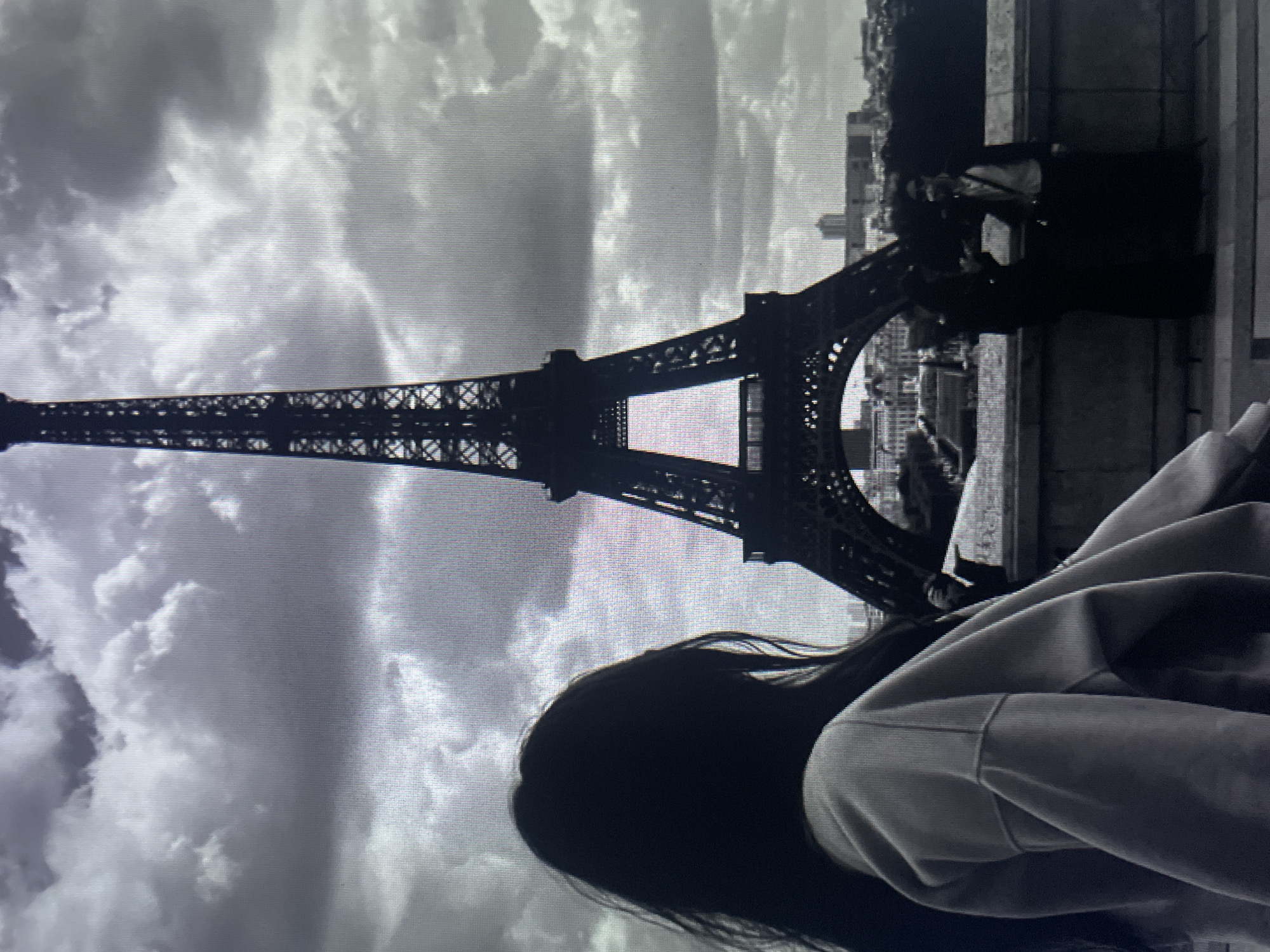 The image is a grayscale, low-resolution photograph featuring the Eiffel Tower in the background under a cloudy sky. In the foreground, the back of a person with long, dark hair is visible, looking towards the tower. They are wearing a light-colored hooded jacket. Further in the background, on a bridge or walkway, two figures are standing. The person on the left is darker and appears to be wearing dark clothing. The person on the right is wearing lighter clothing, possibly a jacket over a shirt, and has glasses. The Eiffel Tower dominates the center of the frame. The city skyline of Paris is visible behind the tower. The photograph does not depict any elements related to Kołobrzeg, Poland.