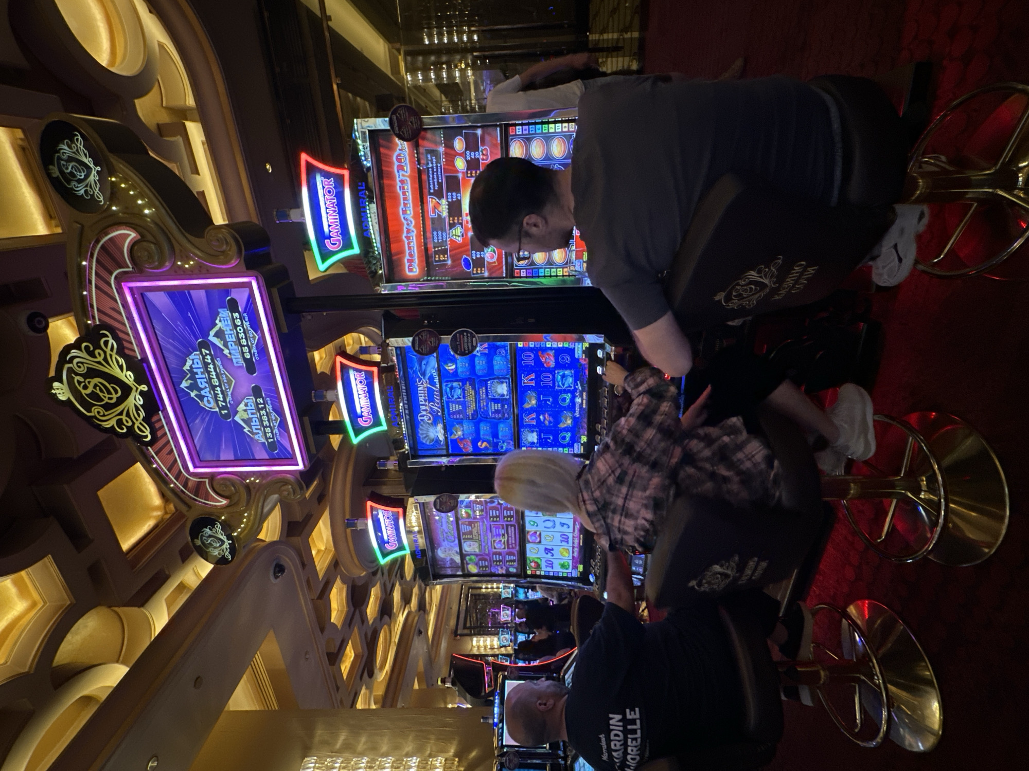 In the city of Estosadok, Russia, at the Casino Sochi, two individuals are seated at slot machines. The person on the left, a bald male wearing a black t-shirt, is facing away from the camera, looking towards a slot machine. To his right, a blonde-haired female with a checkered shirt is also facing a slot machine. Both are seated on gold-colored bar stools. The slot machines are illuminated with colorful lights and display various game graphics and numbers. The ceiling above is ornately decorated with gold accents and recessed lighting, characteristic of a luxurious casino environment. In the background, other slot machines and patrons are visible, creating a lively atmosphere. The floor is covered in a deep red carpet.