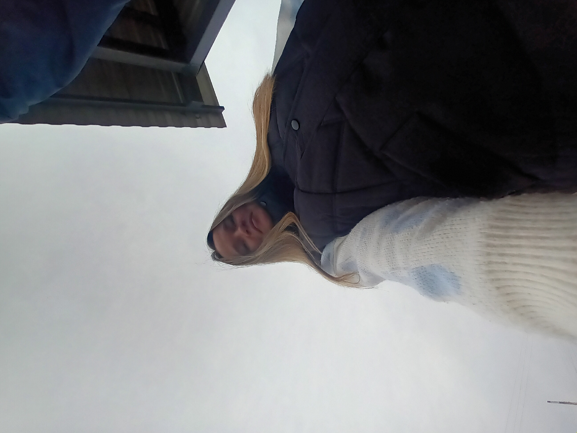 A woman with blonde hair is looking down at the camera. She is wearing a black jacket and a white and blue striped sweater. The sky is grey and overcast. In the upper left corner, there is a building. The location is Ольховка, Russia.