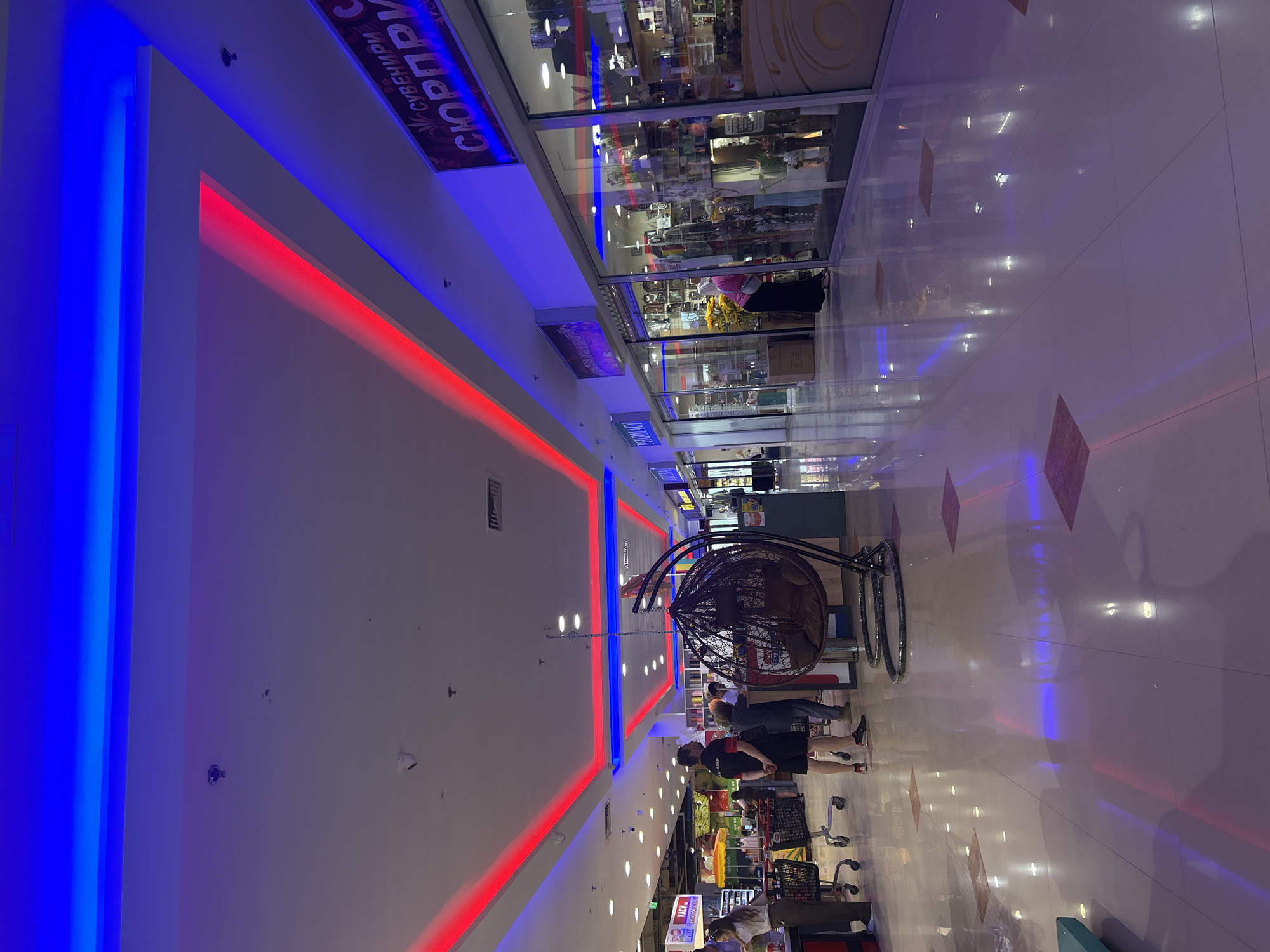 This is a photograph taken inside a modern shopping mall, likely in Shumen, Bulgaria, given the Cyrillic signage. The lighting is dynamic, with prominent blue and red LED strips illuminating the ceiling and the polished floor, creating a vibrant and somewhat futuristic atmosphere. The floor reflects the lights and the surrounding environment, giving it a glossy appearance.
In the foreground, a dark brown, woven hanging chair with plush cushions is a prominent feature, suggesting a seating or relaxation area. To the left, several people are visible. One person, wearing a black shirt and shorts, stands facing away from the camera. Another person, partially visible, is wearing a light-colored top and dark pants, standing near a shopping cart. Further back, there are more shoppers interacting with displays and browsing in the stores.
The shops themselves are set behind glass storefronts, showcasing a variety of merchandise. One store has prominent signage in Cyrillic, with the word "СЮРПРИЗИ" (Surprises) clearly visible, indicating it's a gift or novelty shop. Another sign above a storefront reads "PHARMACY". The stores appear well-stocked with products displayed on shelves.
The overall environment is clean and well-lit, characteristic of a commercial space. The architectural design features clean lines and modern materials. The scene captures a typical day in a shopping mall, with people going about their activities amidst the colorful and ambient lighting.