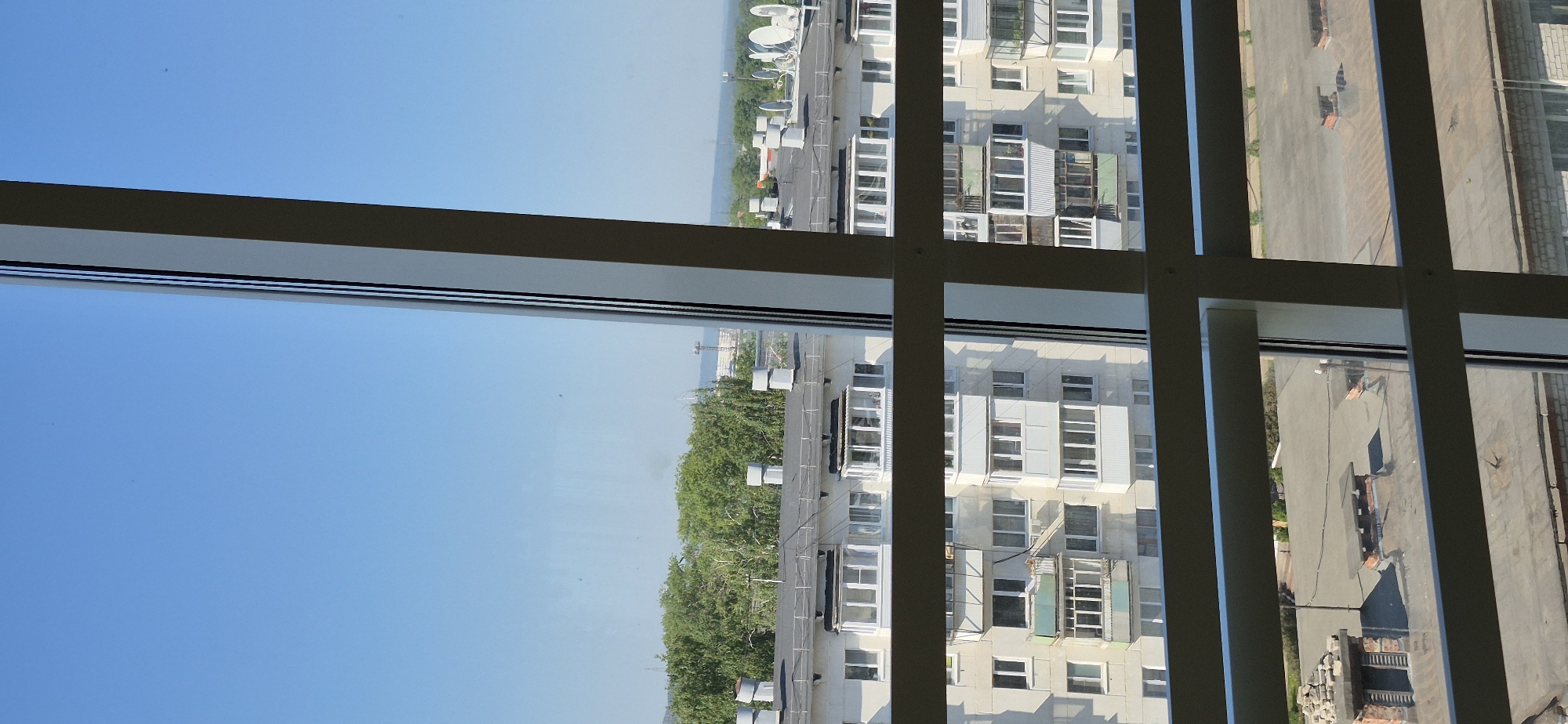 The image depicts a view through a window. The window frame is visible in the foreground, dividing the scene. On the left side of the frame, a clear blue sky is visible, suggesting fair weather. The right side of the frame shows a densely populated urban area, presumably the city of Unknown, Unknown. Several high-rise buildings are present, characterized by numerous windows and balconies. A road and sidewalk are also discernible, with a few indistinct figures that could be people. Trees add a touch of greenery to the cityscape. The overall impression is of a typical urban landscape seen from an elevated viewpoint.
