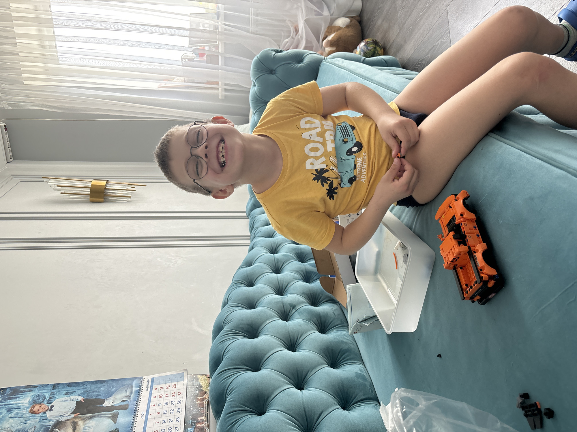 A young boy with glasses is lying on a turquoise tufted couch. He is wearing a yellow shirt with a blue car and palm trees on it. He is smiling and holding orange Lego pieces in his hands. Next to him on the couch is a white container and an orange Lego vehicle. There is a calendar hanging on the wall behind the couch. The room is brightly lit with sunlight coming through the white curtains.