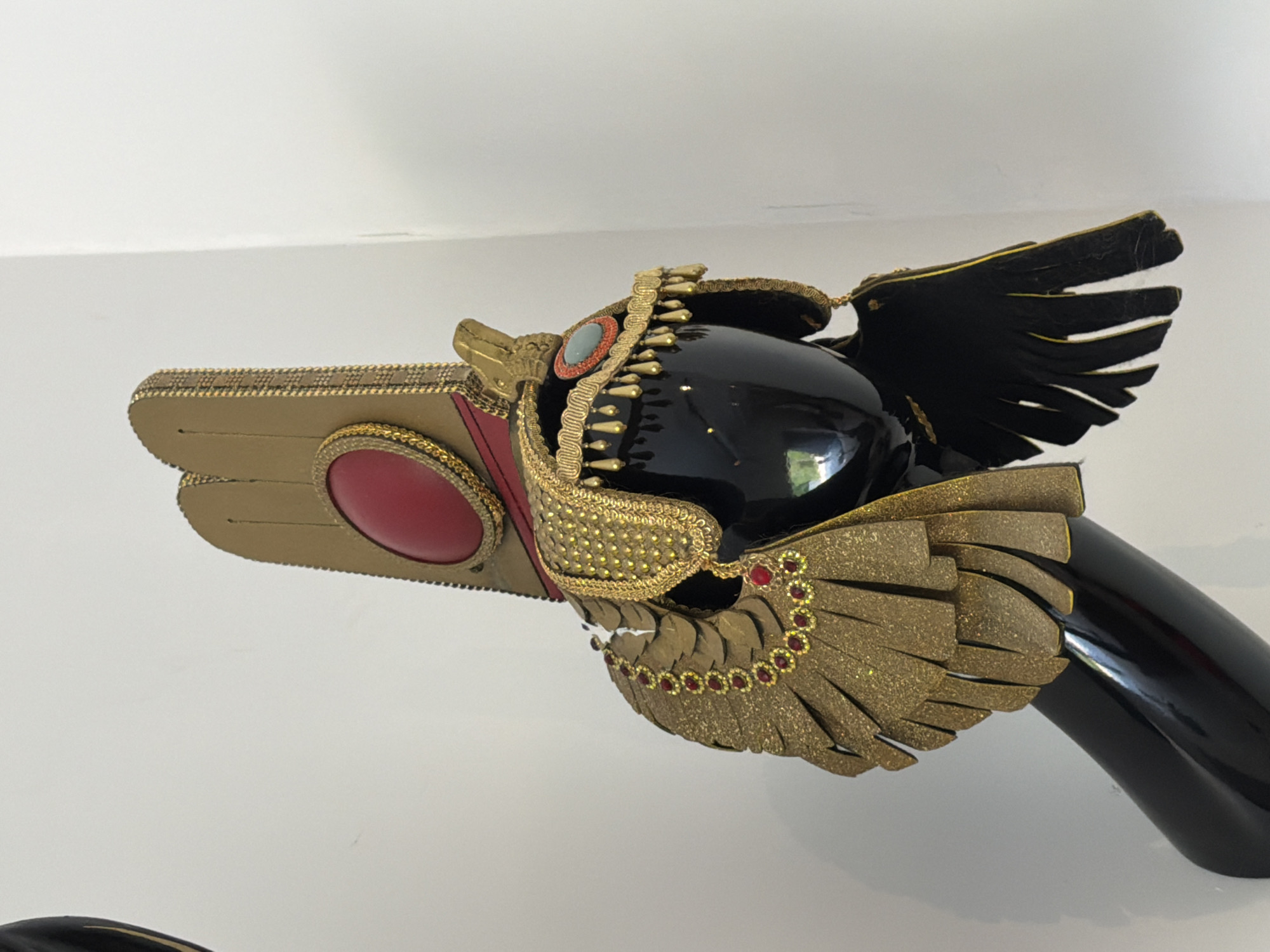 A black mannequin head and bust, tilted slightly to the right, wears an elaborate, golden Egyptian-style headdress. The headdress features a tall, rectangular crown with a large, red, oval-shaped stone set in the center, framed by gold trim. A fringed gold collar adorned with red and gold beads encircles the neck, and stylized gold wings, embellished with red gems, extend from the sides. A smaller, oval, turquoise-colored stone is set into the headdress on the right side. The background is a plain white wall. There are no visible people or explicit signs of location within the city of Ko Samui District, Thailand. The lighting is even, suggesting an indoor setting.