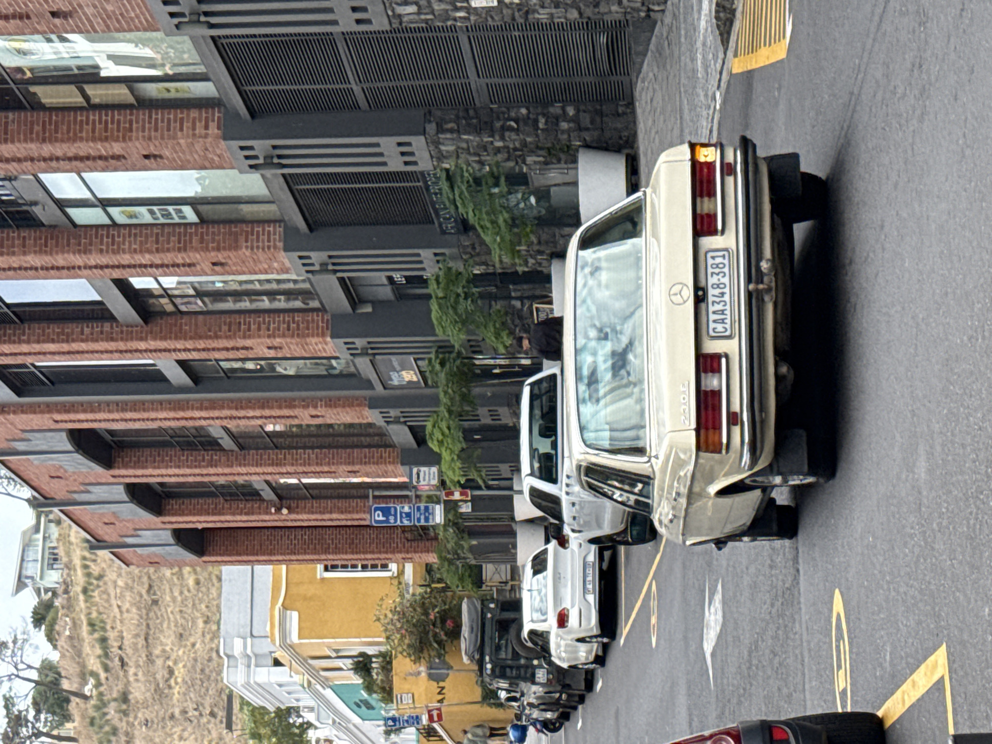 A street scene in Cape Town, South Africa, shows a row of parked vehicles along a street with a slight incline. The vehicles consist of various cars, including a cream-colored older Mercedes-Benz with the license plate "CAA348-381" clearly visible. The buildings along the street are diverse, with a mix of brick structures, modern black-framed buildings, and colorful facades in yellow and green. Street signage, including parking signs, is visible along the road. Trees and other greenery are interspersed along the street. A person is walking on the sidewalk next to the parked cars. The road is marked with yellow lines delineating parking spaces.
