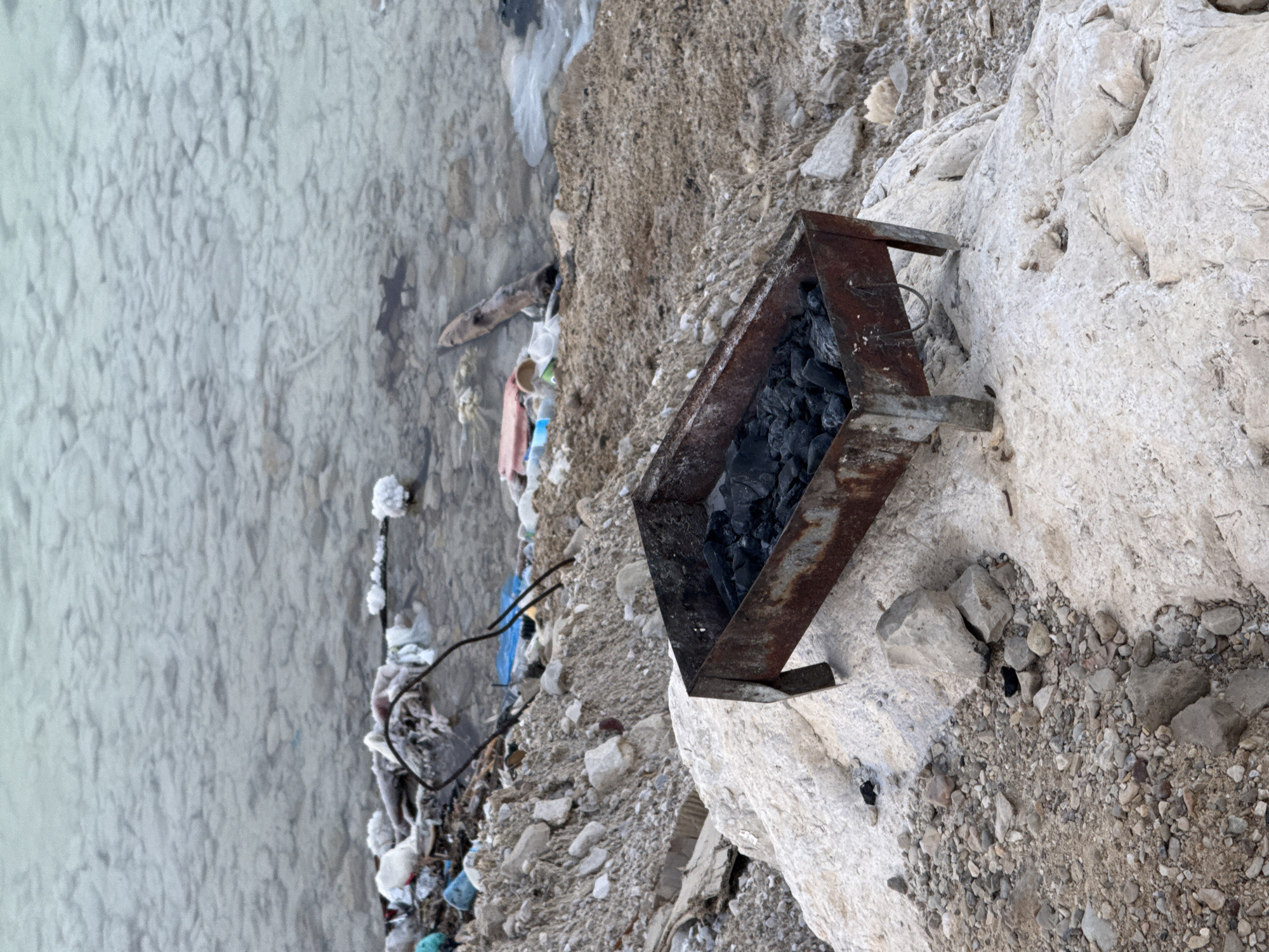 A rusty, rectangular metal grill filled with charcoal sits on a large, light-colored rock near the shore of a body of water. The water is shallow and murky, with a rocky bottom visible. The shoreline is littered with various debris, including plastic bags, bottles, and other refuse. The background consists of a sandy, rocky slope leading away from the water. The scene suggests a neglected or abandoned area, possibly a picnic spot that has fallen into disrepair. The location in the city of Tamar Regional Council, Israel is consistent with such a scene, as this region is known for its diverse natural landscapes and potential for outdoor recreation, but also faces challenges with litter and pollution.