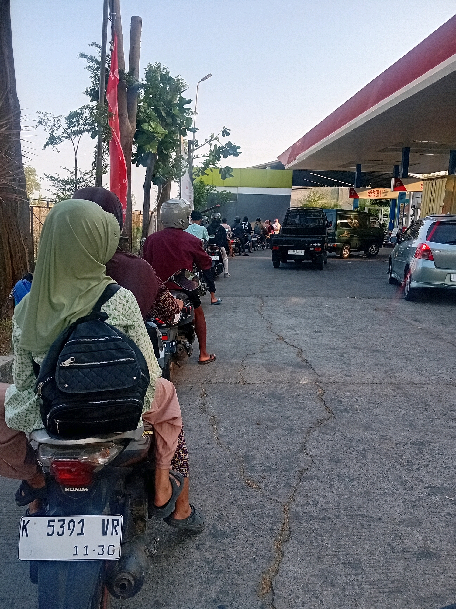 The image depicts a queue of motorcycles and cars at a gas station in Bacin, Indonesia. In the foreground, a woman wearing a light green hijab and patterned shirt rides on the back of a motorcycle, with a black backpack strapped to her. Her legs are visible, wearing light brown pants and dark sandals. The motorcycle has a license plate reading "K 5391 VR" and "11.30". Behind her, other motorcycles and their riders are also waiting in line.
The gas station features a red and white canopy covering the pumps. Several vehicles, including a black pickup truck and a green van, are visible further down the queue. The ground is asphalt with visible cracks. In the background, trees and buildings are present. The sky is a clear, pale blue, suggesting it is daytime. A red flag is seen attached to a wooden pole on the left side of the image.