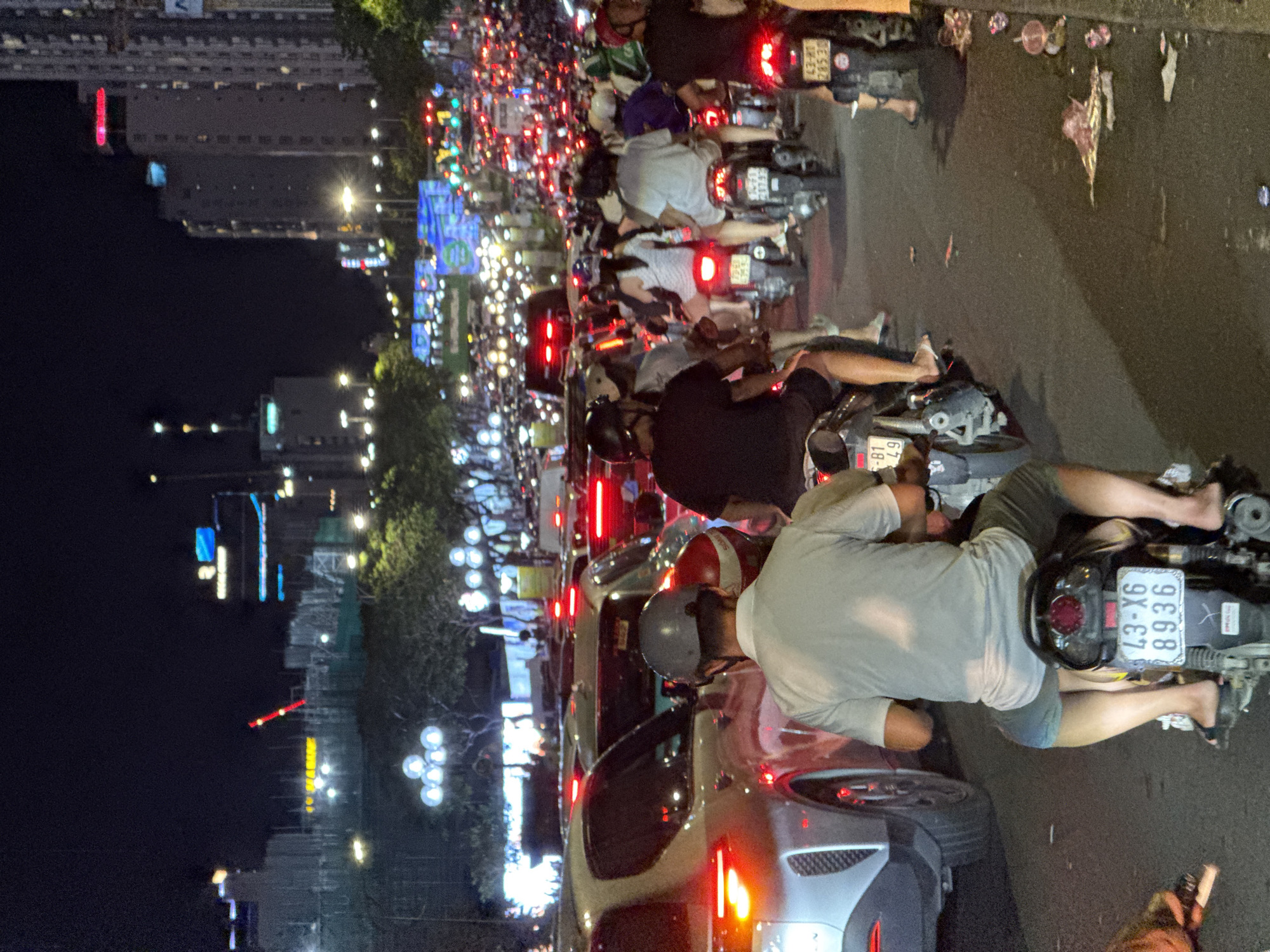 This image captures a night scene of severe urban traffic congestion in Đà Nẵng, Vietnam. The foreground is dominated by the back of a male motorcyclist, wearing a grey helmet, a light grey short-sleeved shirt, and dark shorts, seen from behind. He is barefoot and resting his feet on the ground or pegs. His black motorcycle bears the license plate "43-X6 8936." To the immediate left, the rear quarter of a light-colored, modern SUV with illuminated red taillights is visible, stopped in traffic.
The road stretches forward into a dense mass of vehicles, primarily motorcycles and cars, all displaying illuminated red taillights, indicating slow or stopped movement. Numerous individuals, mostly wearing helmets, are visible on their motorcycles, with some carrying passengers. A person on a motorcycle on the far right wears a distinctive green and white helmet, characteristic of the Grab ride-hailing service.
In the mid-ground, large, illuminated billboards or screens with blue and green graphics are visible above the traffic. The background features several multi-story buildings, some with illuminated windows and signage, including a prominent red construction crane on the horizon to the left. Streetlights illuminate the scene from above. The asphalt road surface on the right side of the frame, near the curb, shows scattered litter and debris. The overall impression is a bustling city experiencing a peak traffic period.
